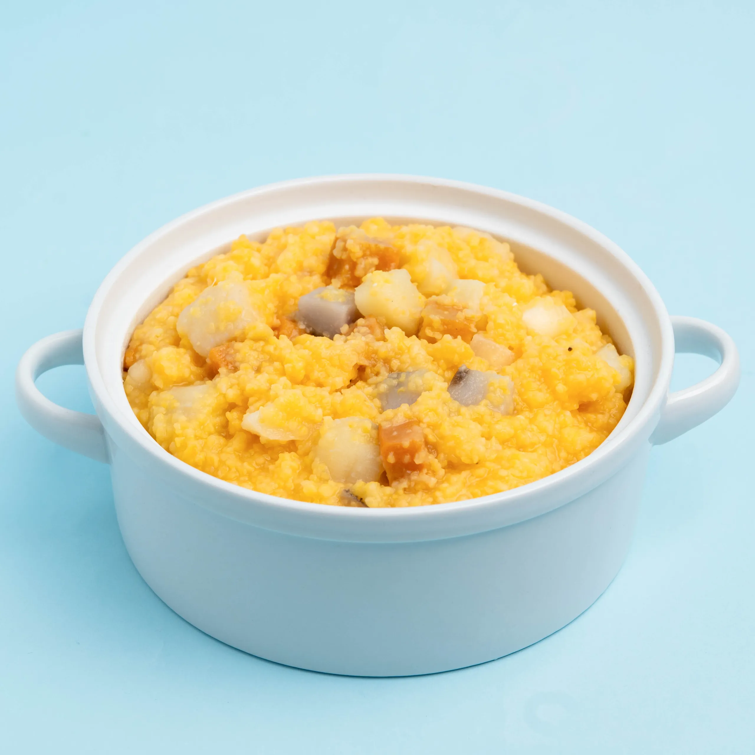 Close-up of a white pot filled with a creamy orange cheese dish containing visible potato chunks and mushrooms, set against a pastel blue background.
