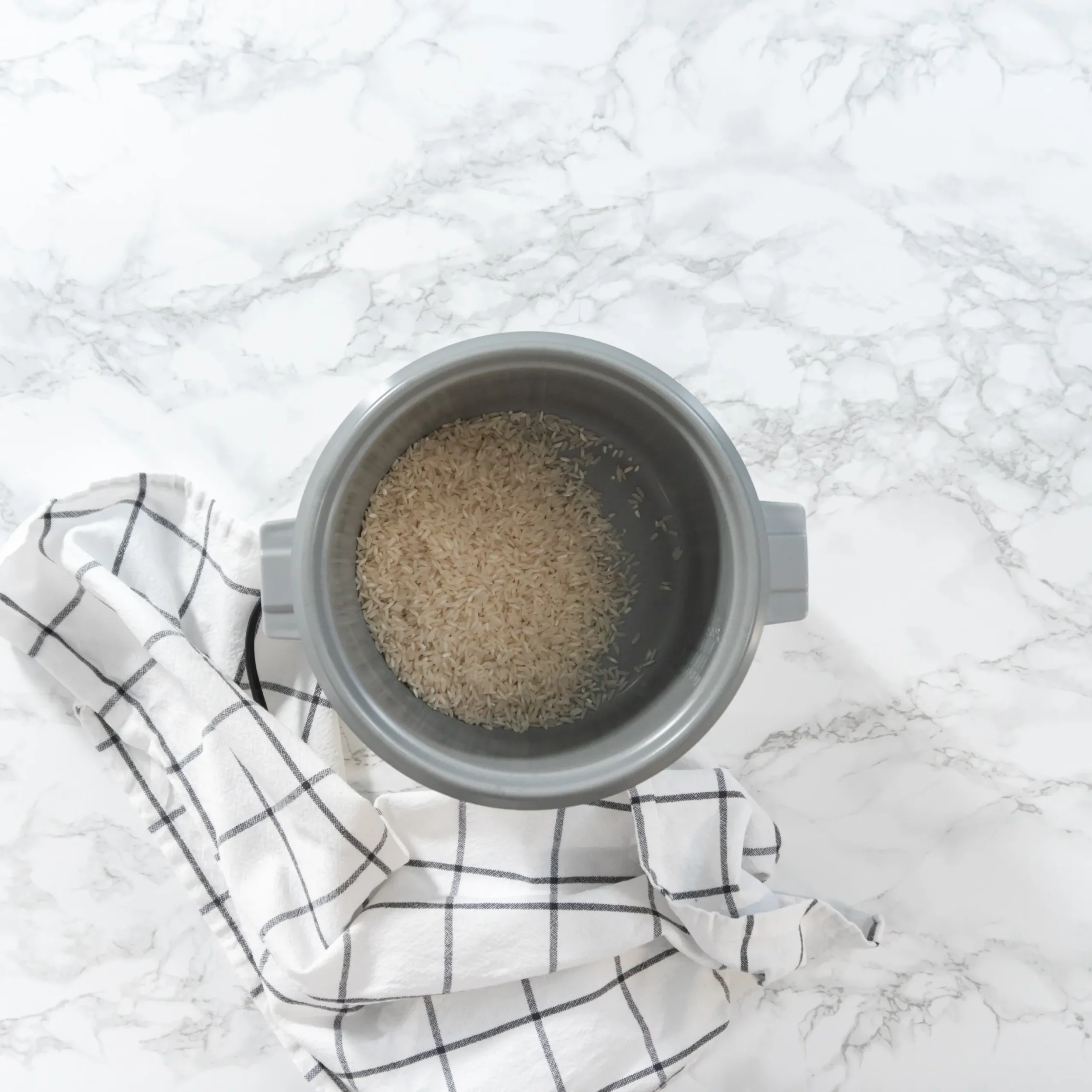 Top-down view of a rice cooker filled with light-brown rice on a white marble countertop, with a striped towel nearby