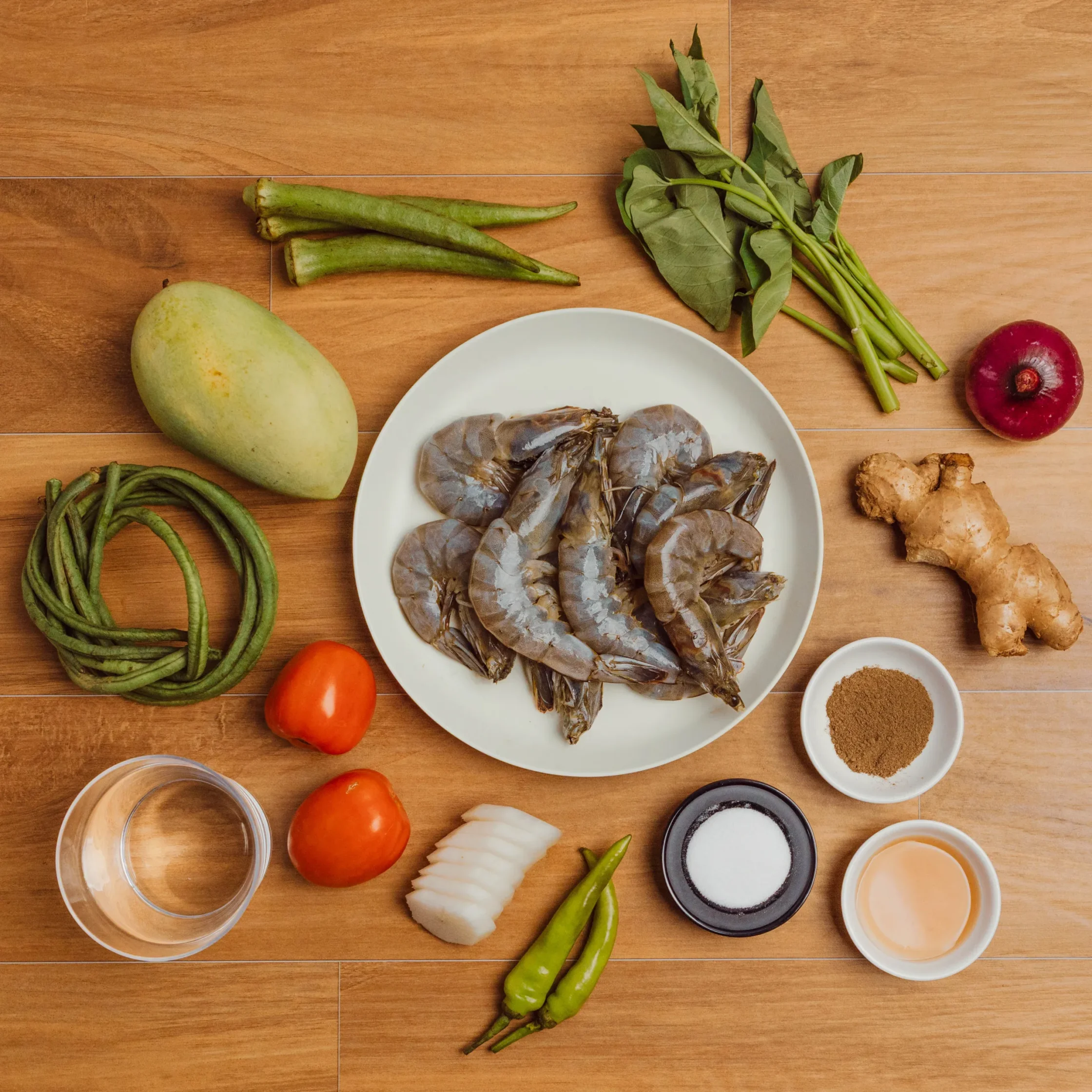 White plate piled with raw shrimp in shells, surrounded by produce on a wooden table (green beans, mango, tomato, onion, ginger) and small bowls of spices.