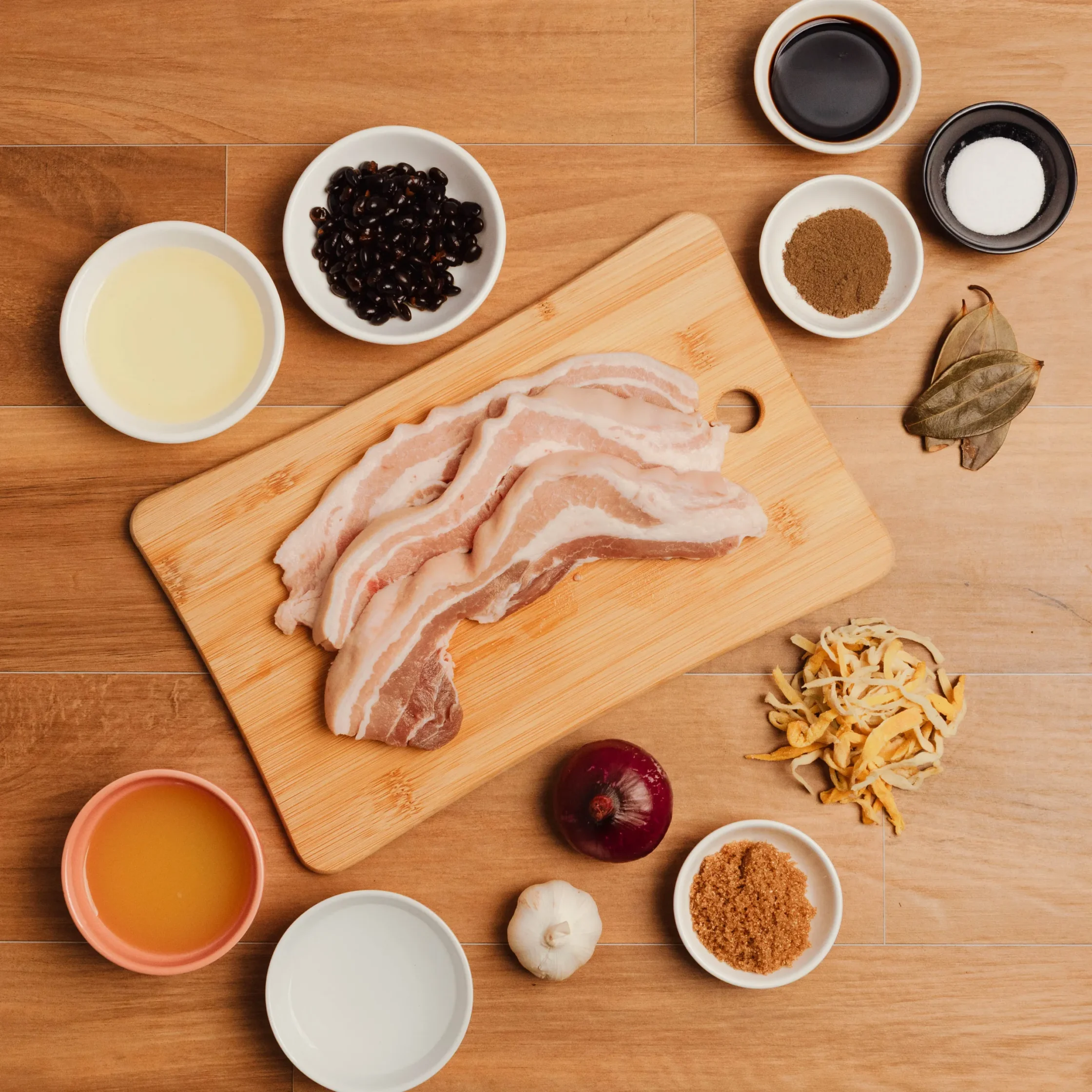 Raw bacon slices arranged on a wooden cutting board with bowls of sauce, brown sugar, and other ingredients nearby on a wooden counter, as part of a recipe setup.