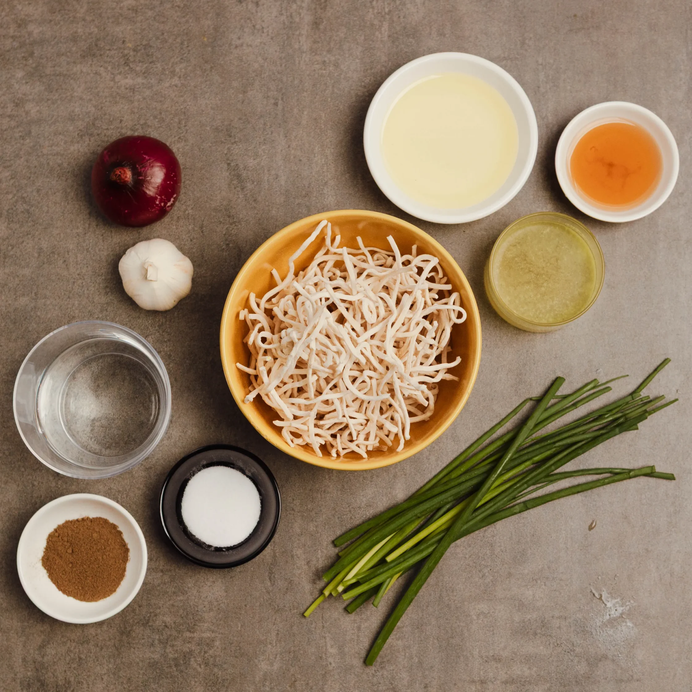 Top-down view of cooking ingredients: a bowl of shredded noodles surrounded by garlic, onion, spices, sauces, and fresh green onions on a grey countertop.