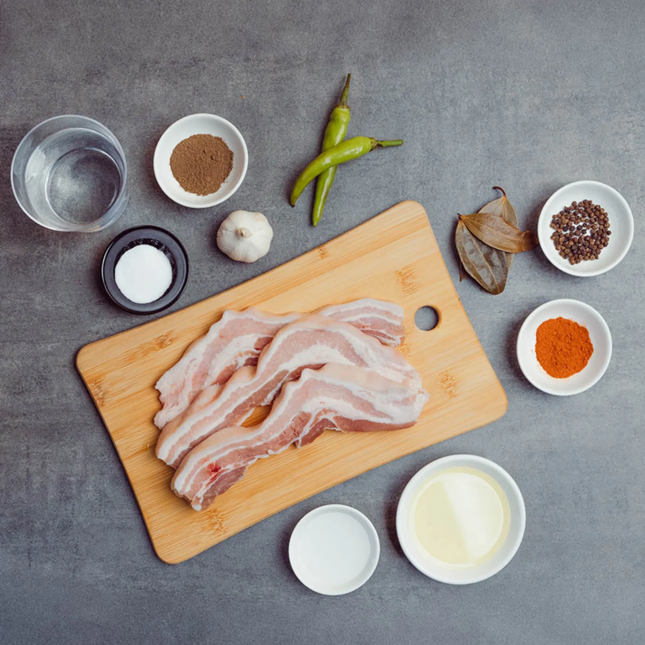 Flat lay of cooking ingredients: bacon slices on a wooden board with bowls of spices, garlic, peppers, and salt on a gray surface