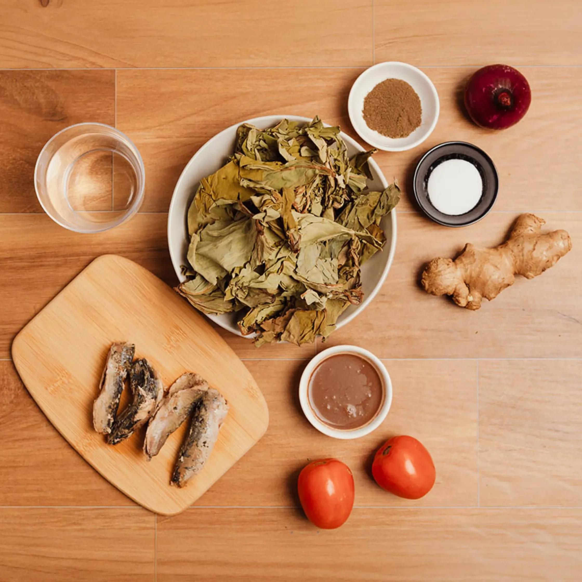 Top-down view of a kitchen counter: a cutting board with a few pieces of grilled fish, fresh tomatoes, a glass of water, a bowl of dried leaves, and small bowls containing brown sauce and a powder on a wooden surface.