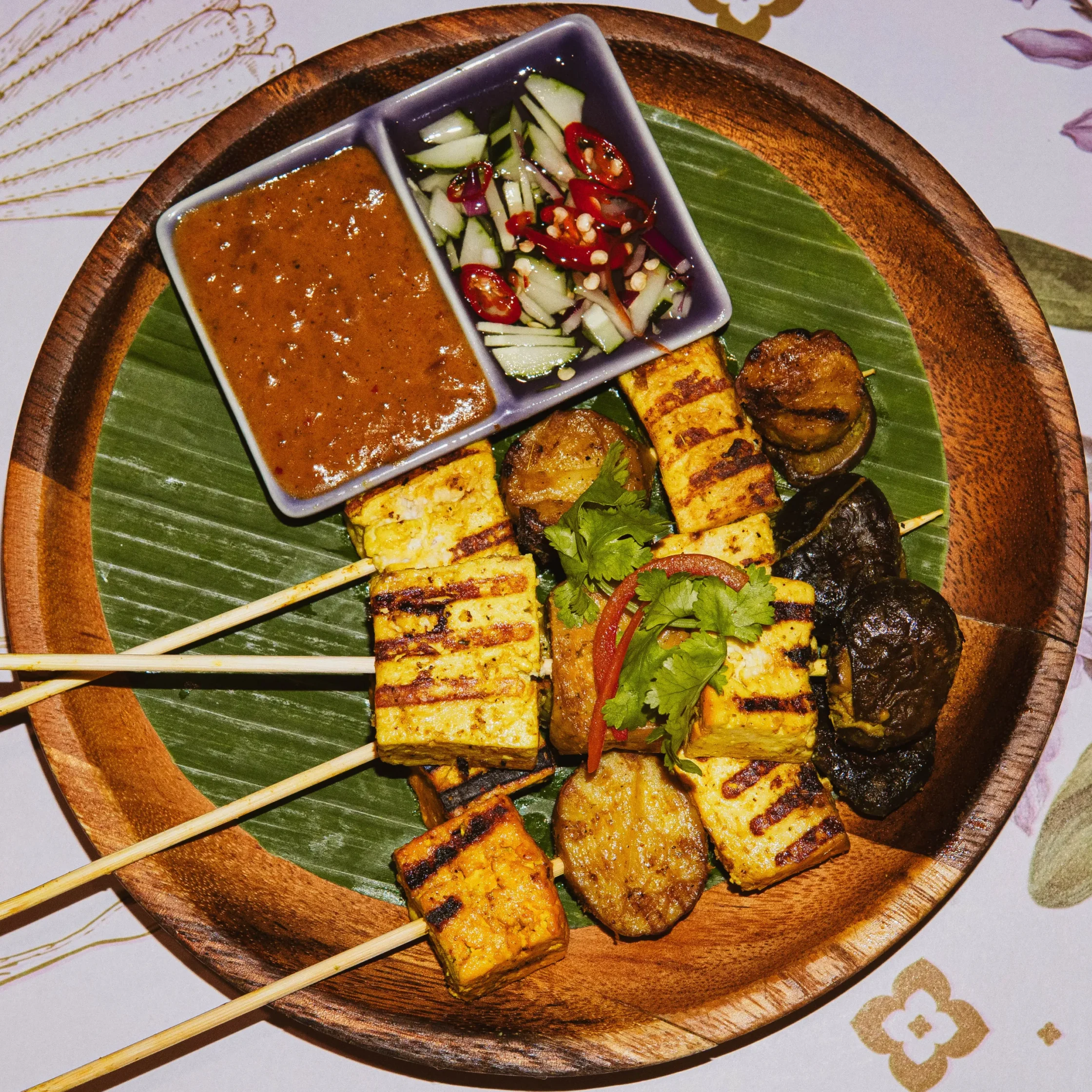 Grilled tofu skewers with a brown dipping sauce and cucumber-red chili salad on a banana-leaf-lined wooden platter