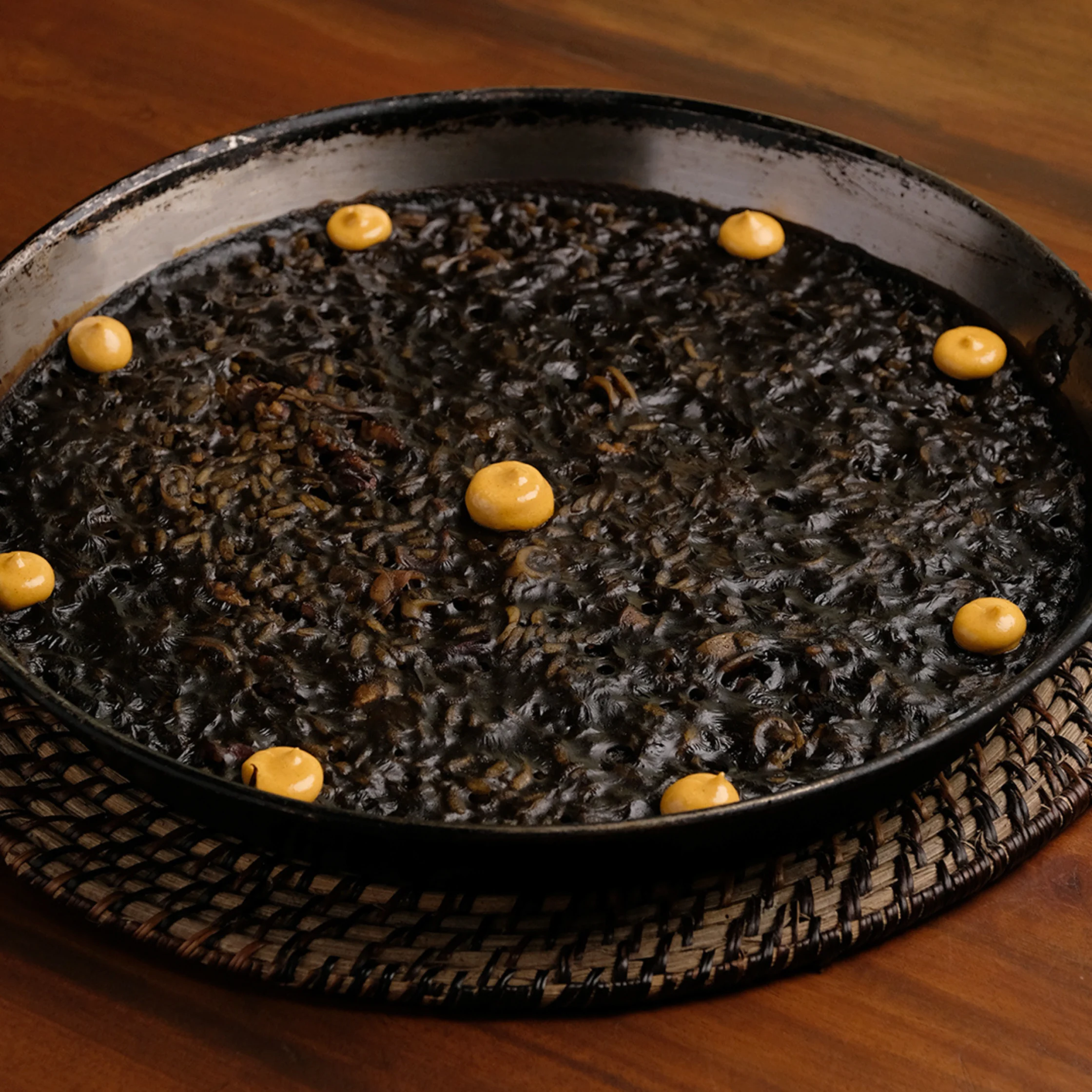Overhead view of a rustic black pan filled with glossy black rice, dotted with golden sauce, on a woven trivet over a wooden table.