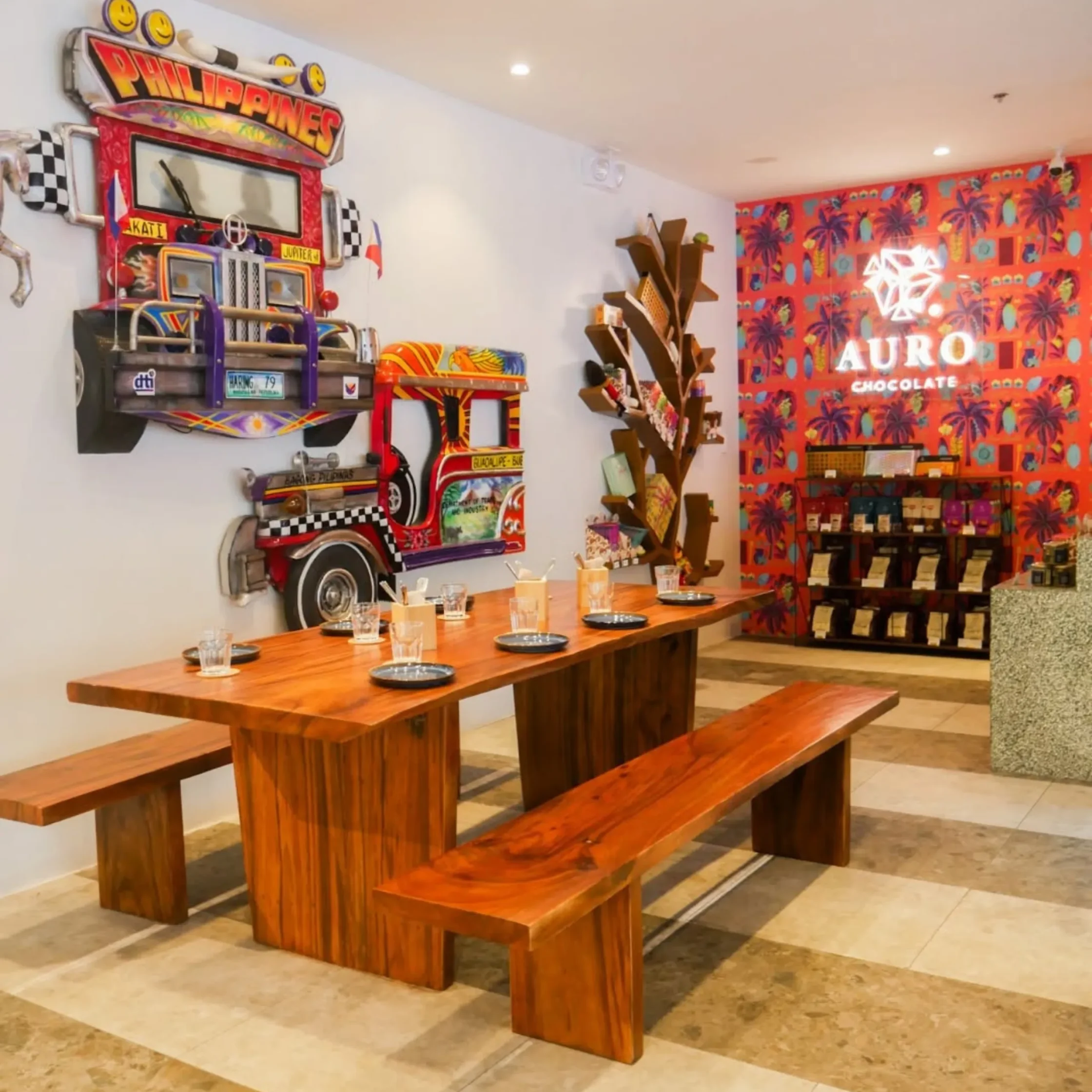Cafe interior with a large retro vehicle sculpture mounted on the wall, wooden communal tables, and a bright palm-pattern mural on the wall behind.