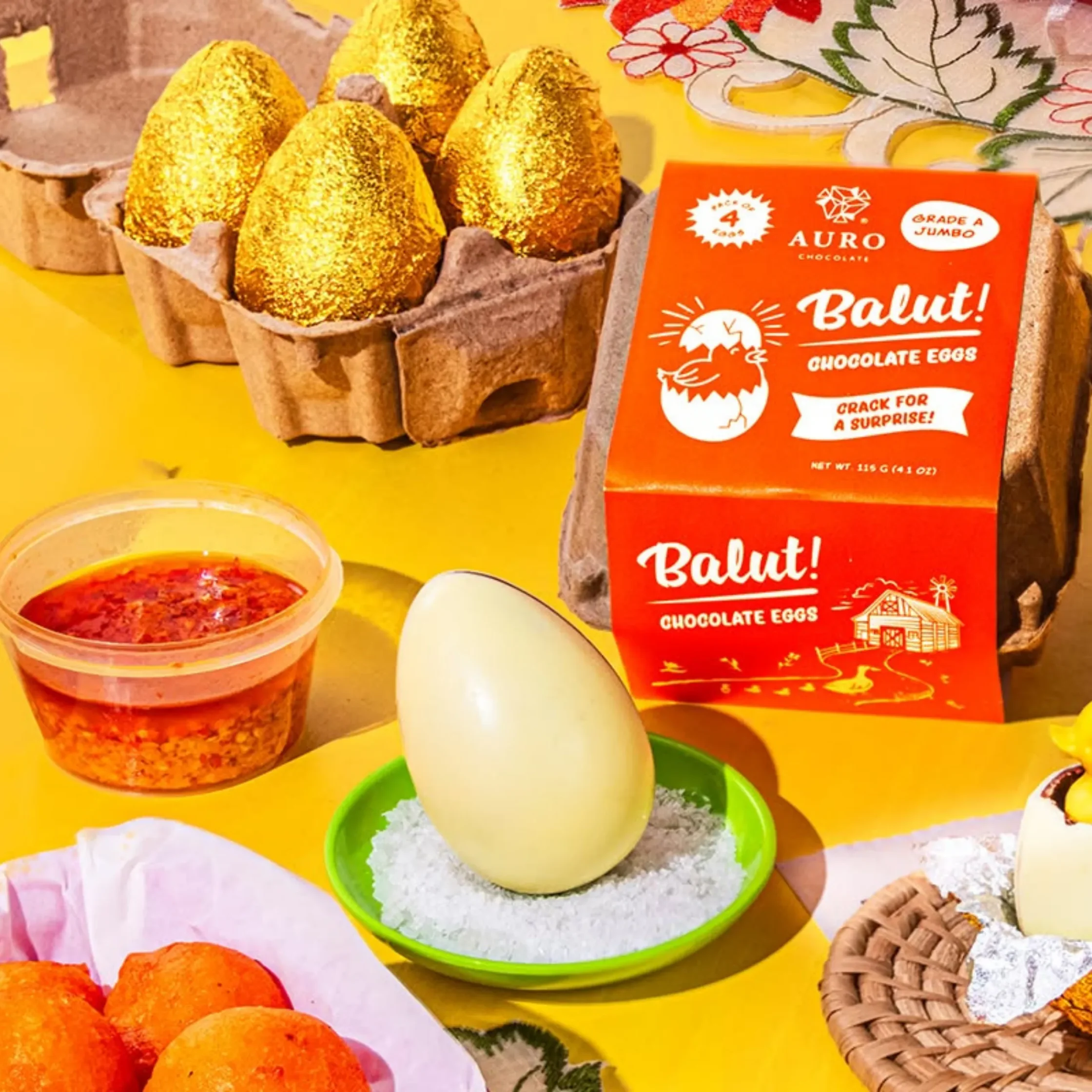 Gold foil-wrapped chocolate eggs in paper cups beside an orange Balut! chocolate eggs box on a bright yellow background.