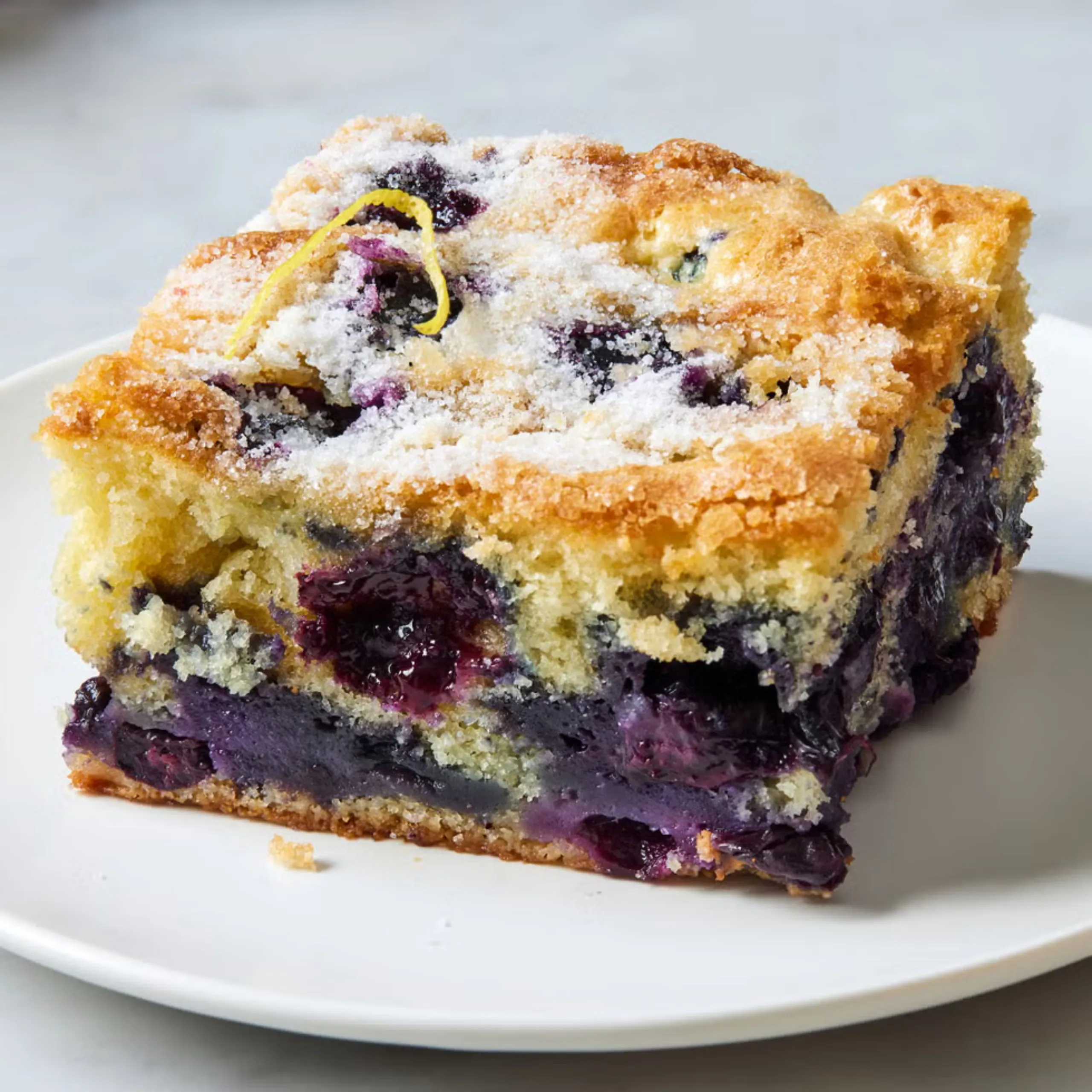 Close-up of a blueberry lemon crumb cake slice on a white plate, dusted with sugar crystals and a lemon zest curl on top.