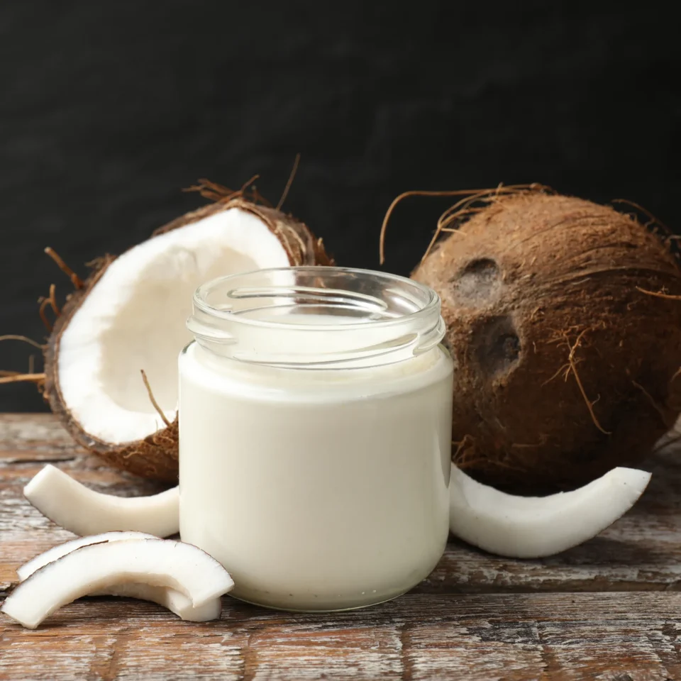 Glass jar of coconut milk with coconut halves and shards on a rustic wooden surface.