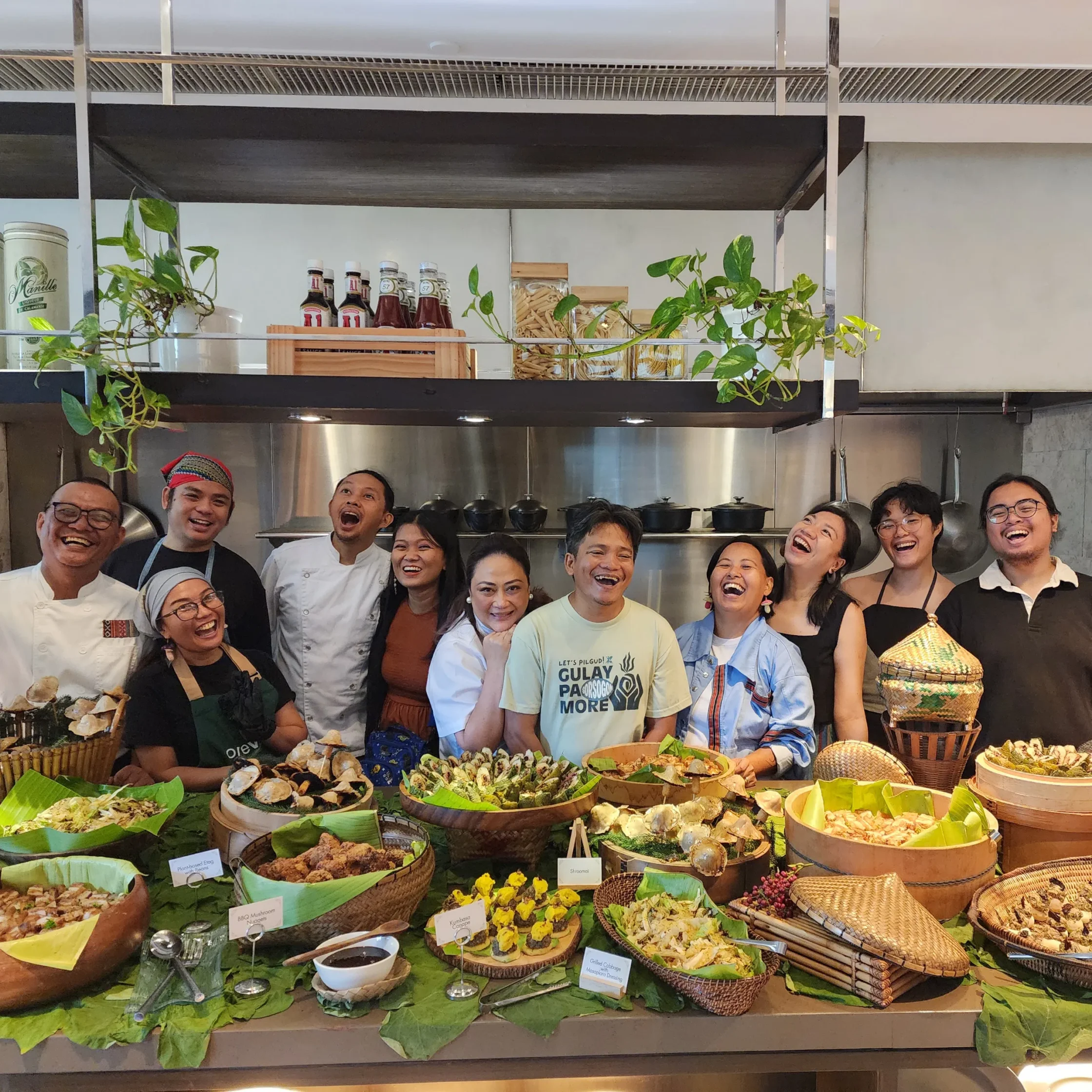 Group of people smiling in a market kitchen behind baskets of assorted foods and greenery on display shelves.