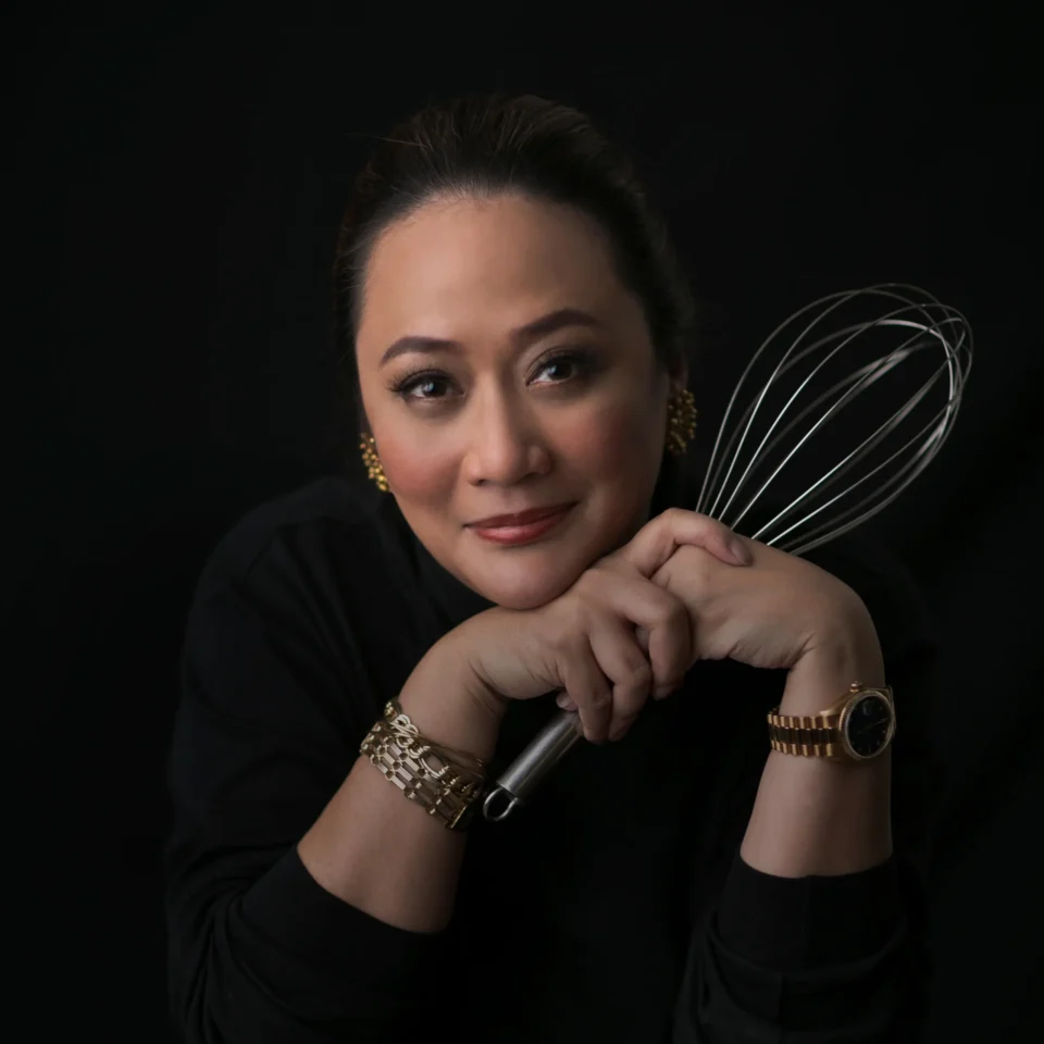 Close-up portrait of a woman smiling while holding a metal whisk against a black background.