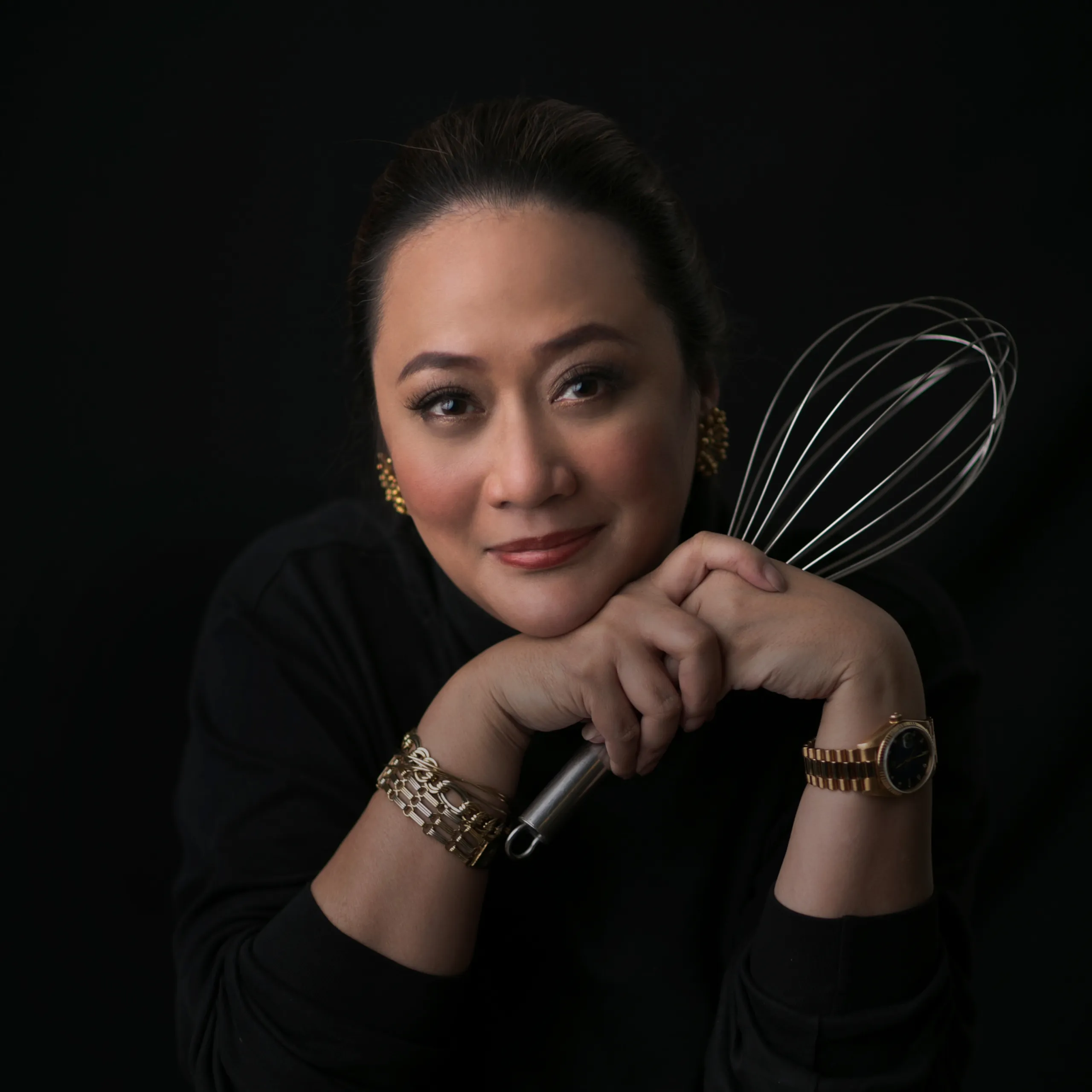 Close-up portrait of a woman smiling while holding a metal whisk against a black background.