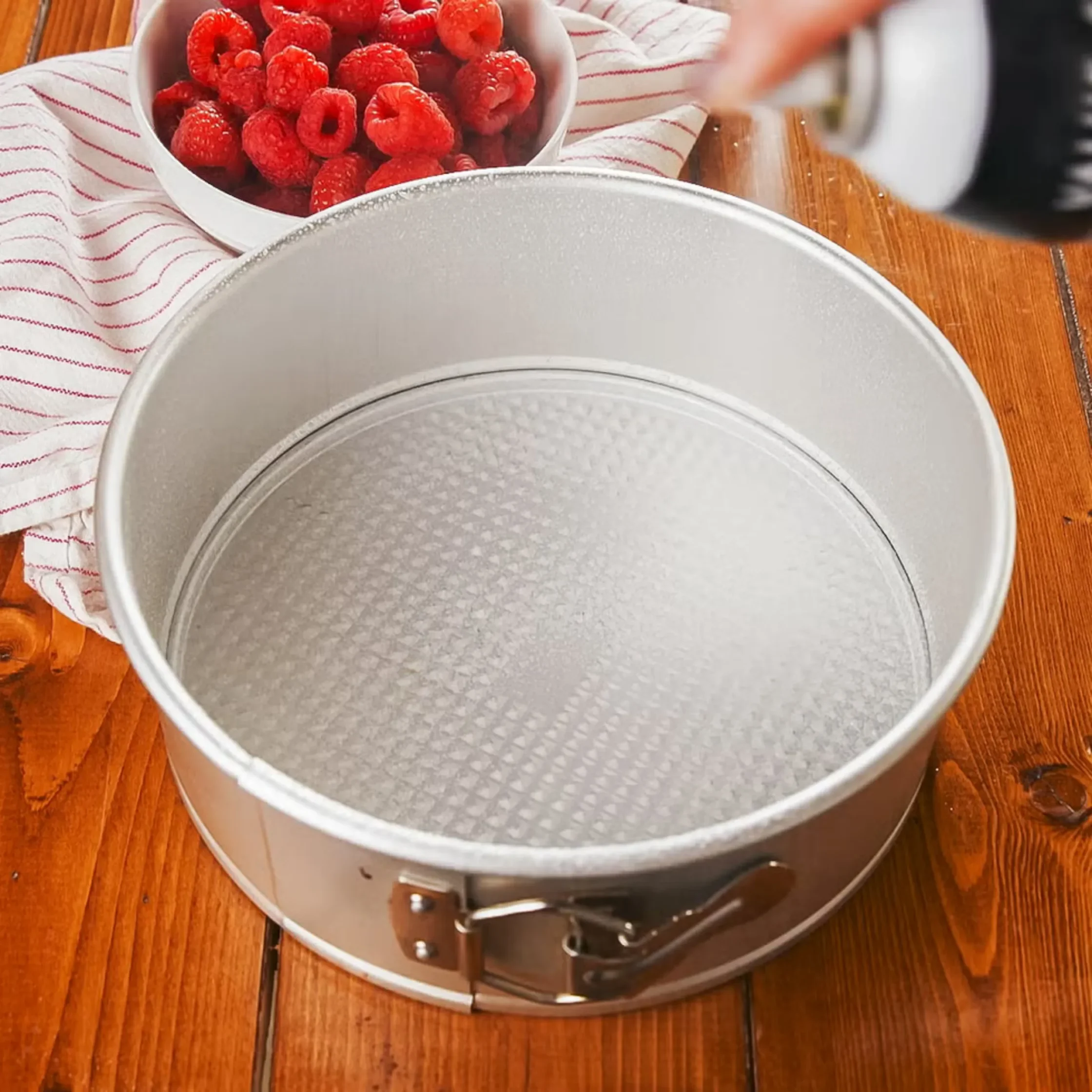 Metal springform cake pan placed on a wooden counter, with a bowl of raspberries and a white towel with red stripes in the background.