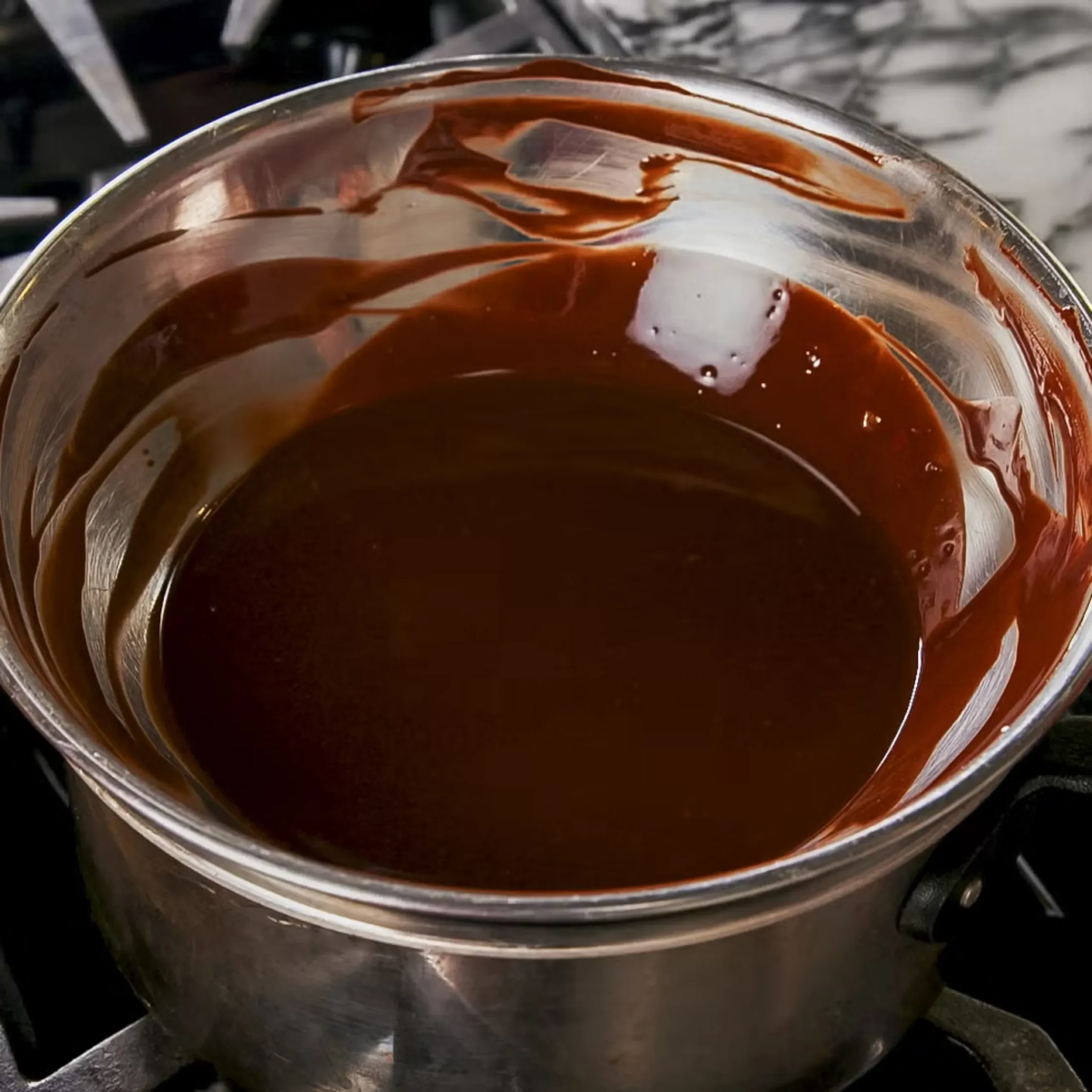 Close-up of a shiny metal pot filled with dark brown liquid, viewed from above, with reflections on the surface and a handle visible nearby.