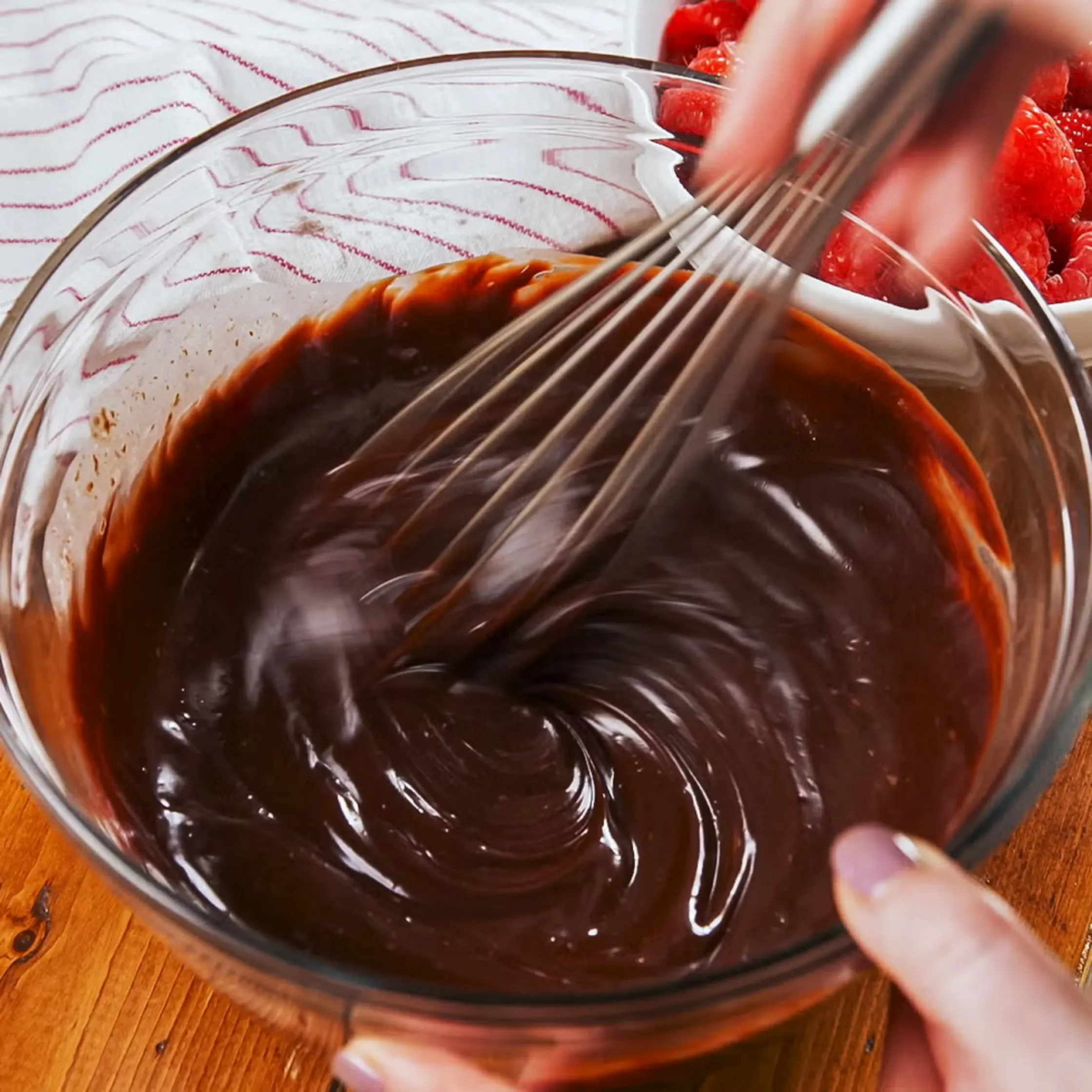 Dark chocolate sauce being whisked in a glass bowl on a wooden table, with red raspberries in the background.