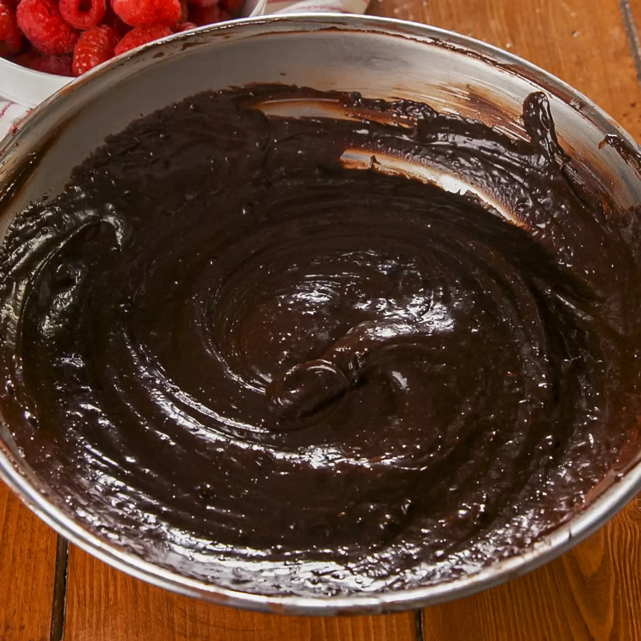 Close-up of glossy chocolate ganache in a metal bowl on a wooden table, with a bowl of raspberries visible in the background.