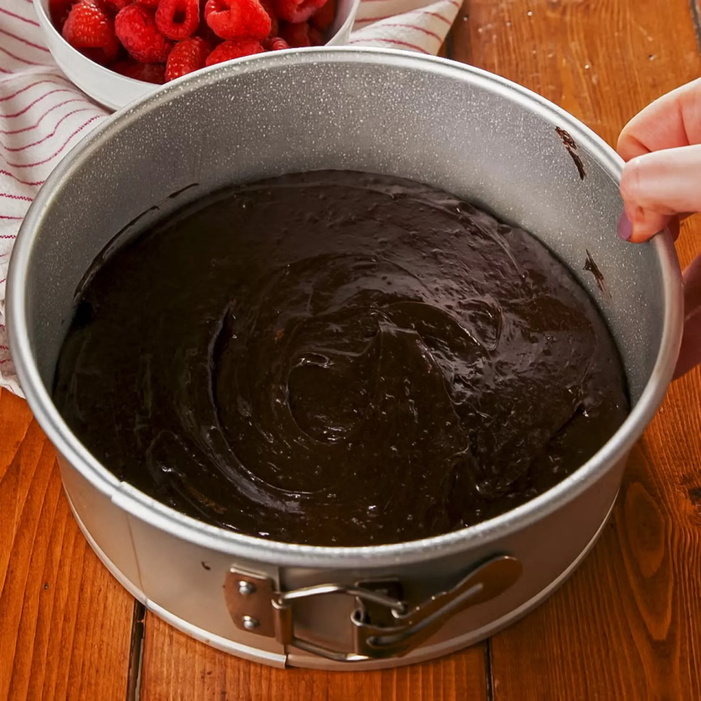 Close-up of a springform pan filled with glossy chocolate batter, with a bowl of raspberries in the background.