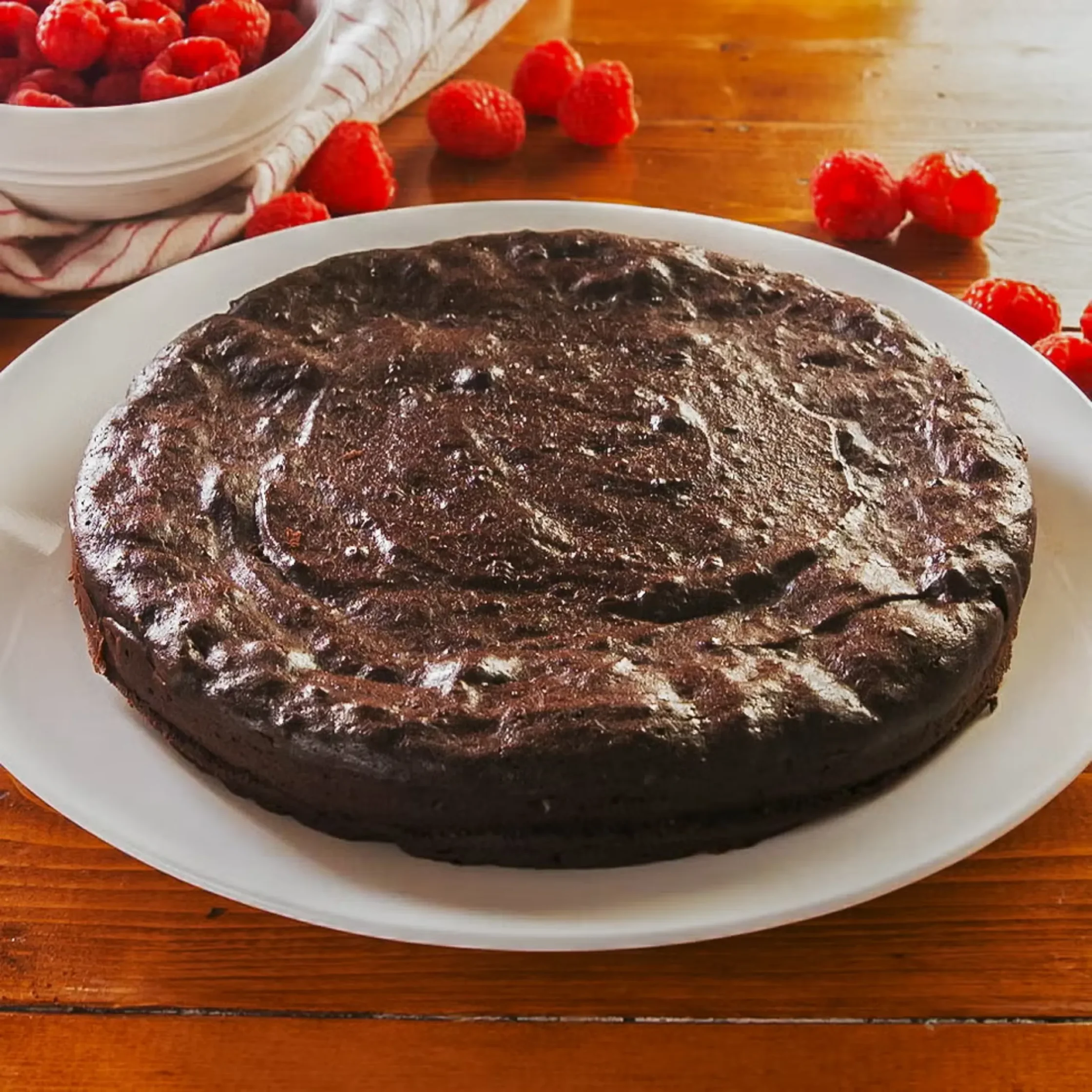 Chocolate cake on a white plate with a light dusting of sugar, raspberries in a bowl nearby on a wooden table