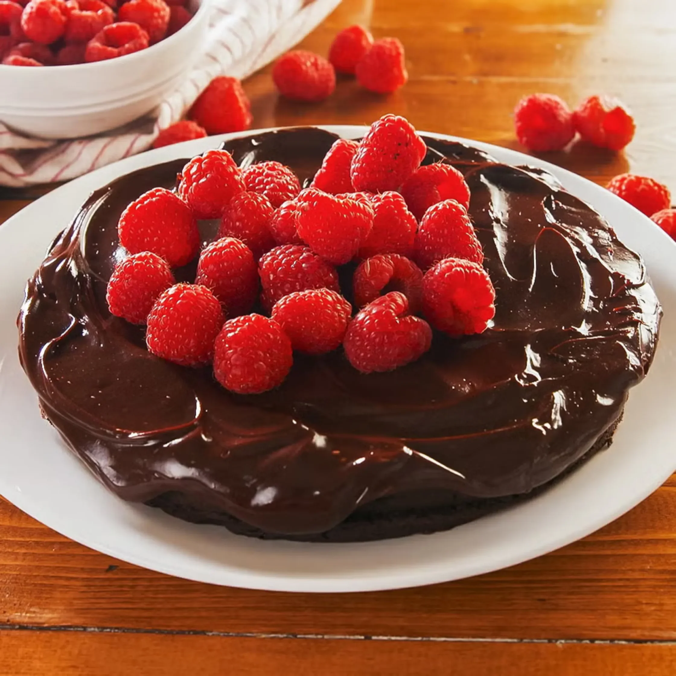 Chocolate cake with glossy ganache topped with fresh raspberries on a white plate