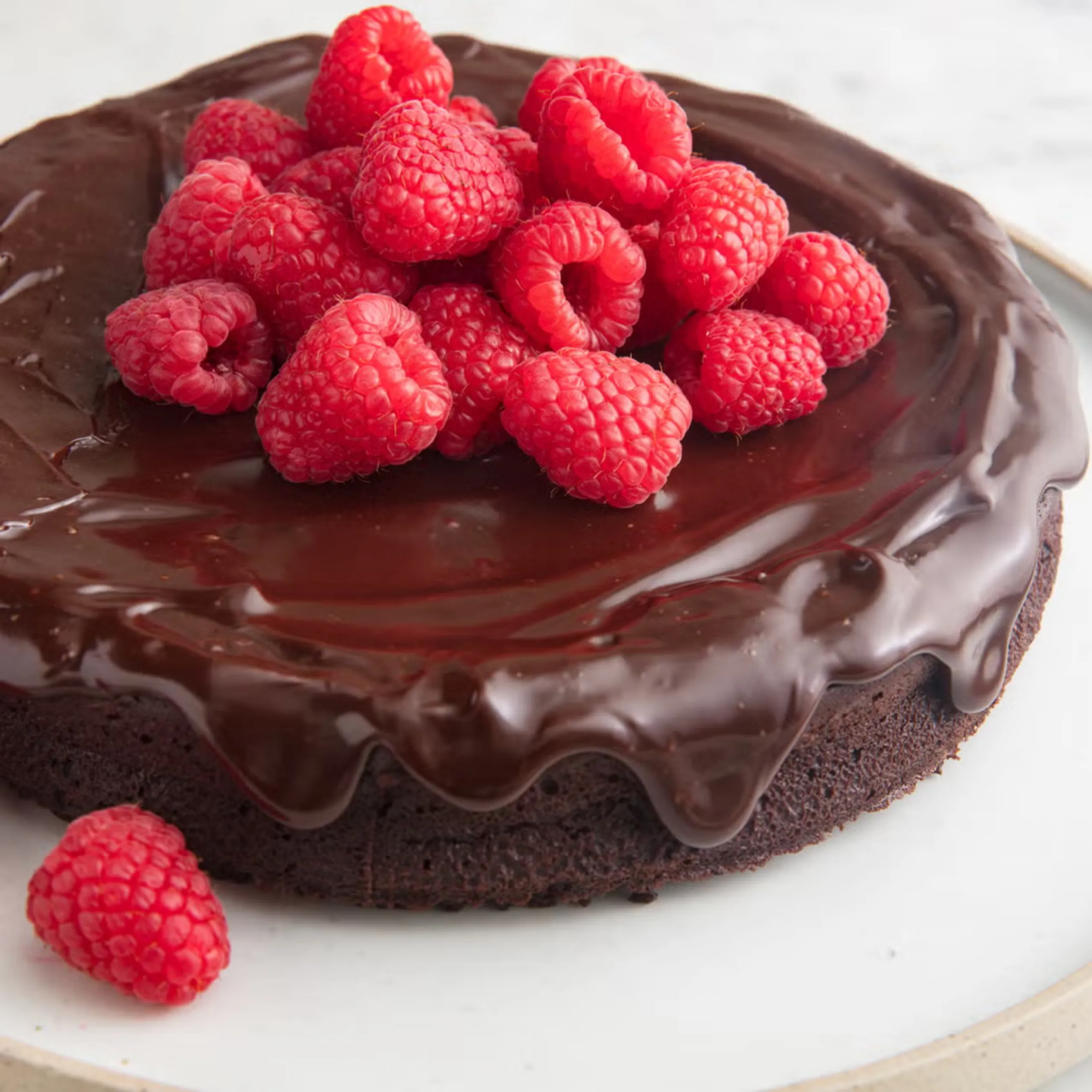 Close-up of fresh red raspberries piled on glossy chocolate ganache topping of a chocolate cake slice on a white plate, highlighting the fruit and dessert.