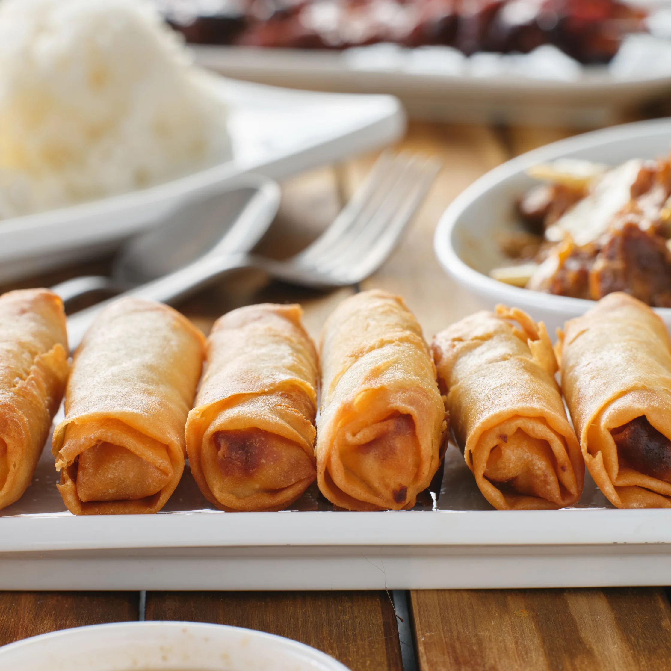 Golden, crispy spring rolls line a white platter, with rice and dipping sauce in the background, creating a warm, inviting meal scene.