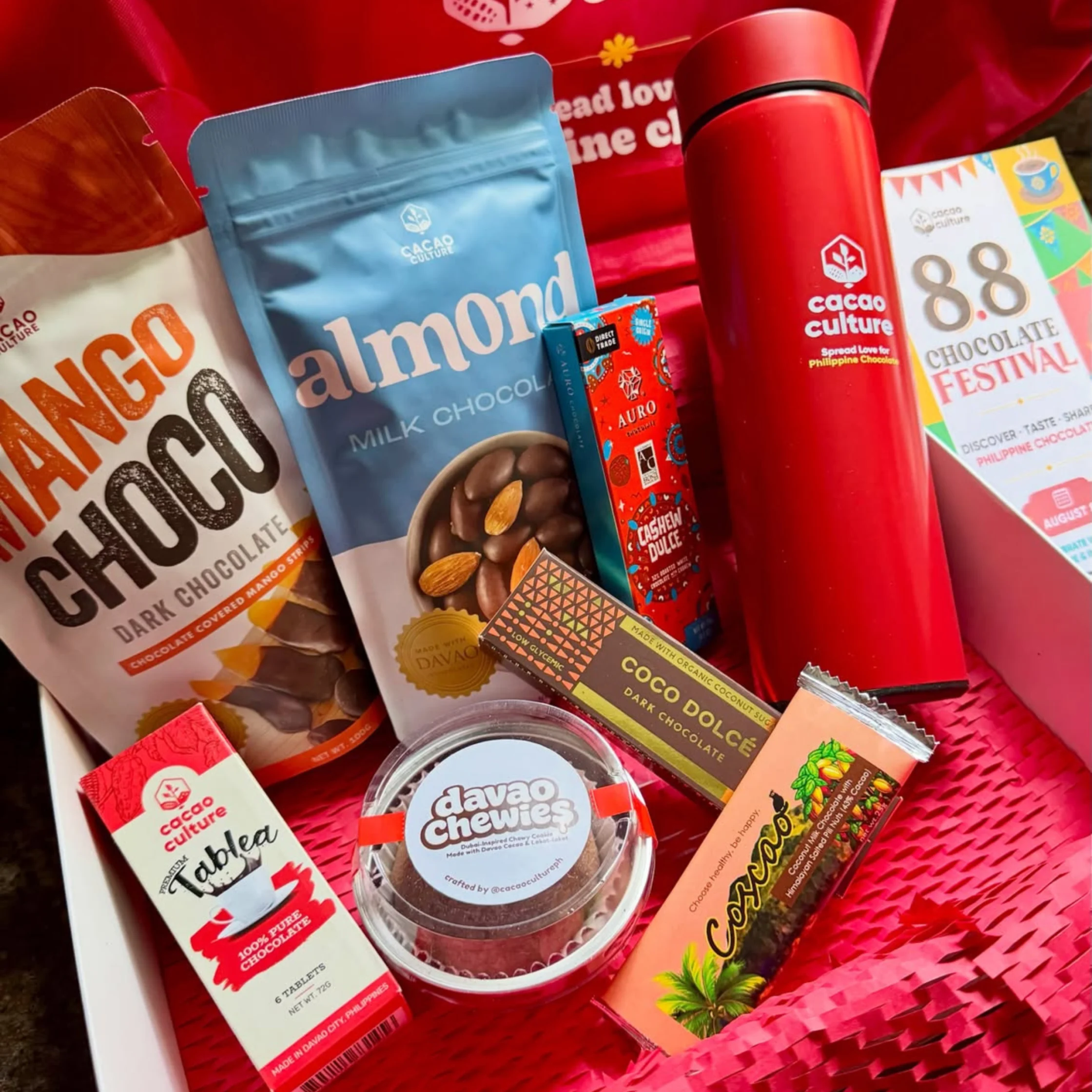 Assorted chocolate bars and treats in a gift box, including Cacao Culture packets and a red bottle-shaped container.