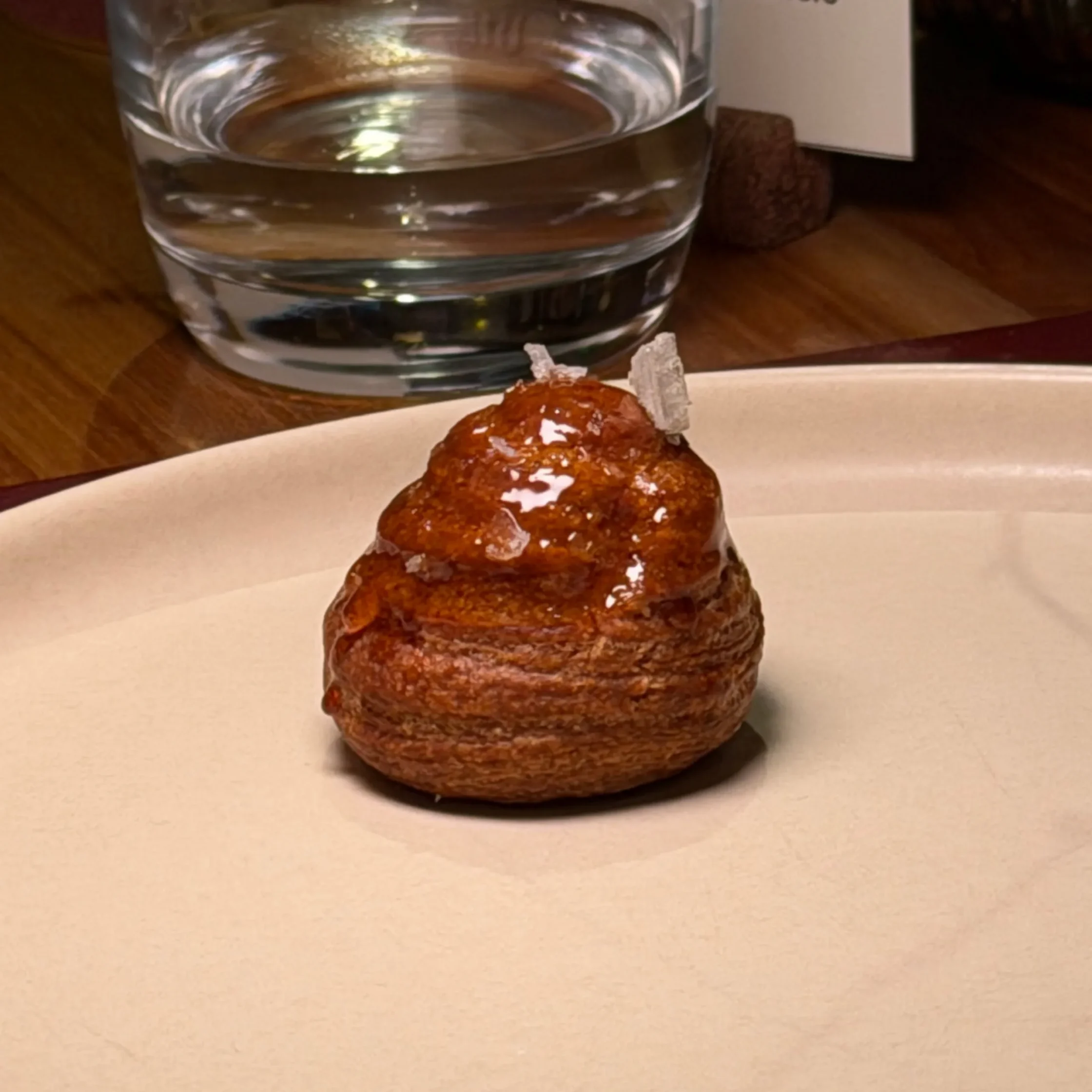 Close-up of a glass of water on a wooden table with a plate of glazed pastries in the foreground.