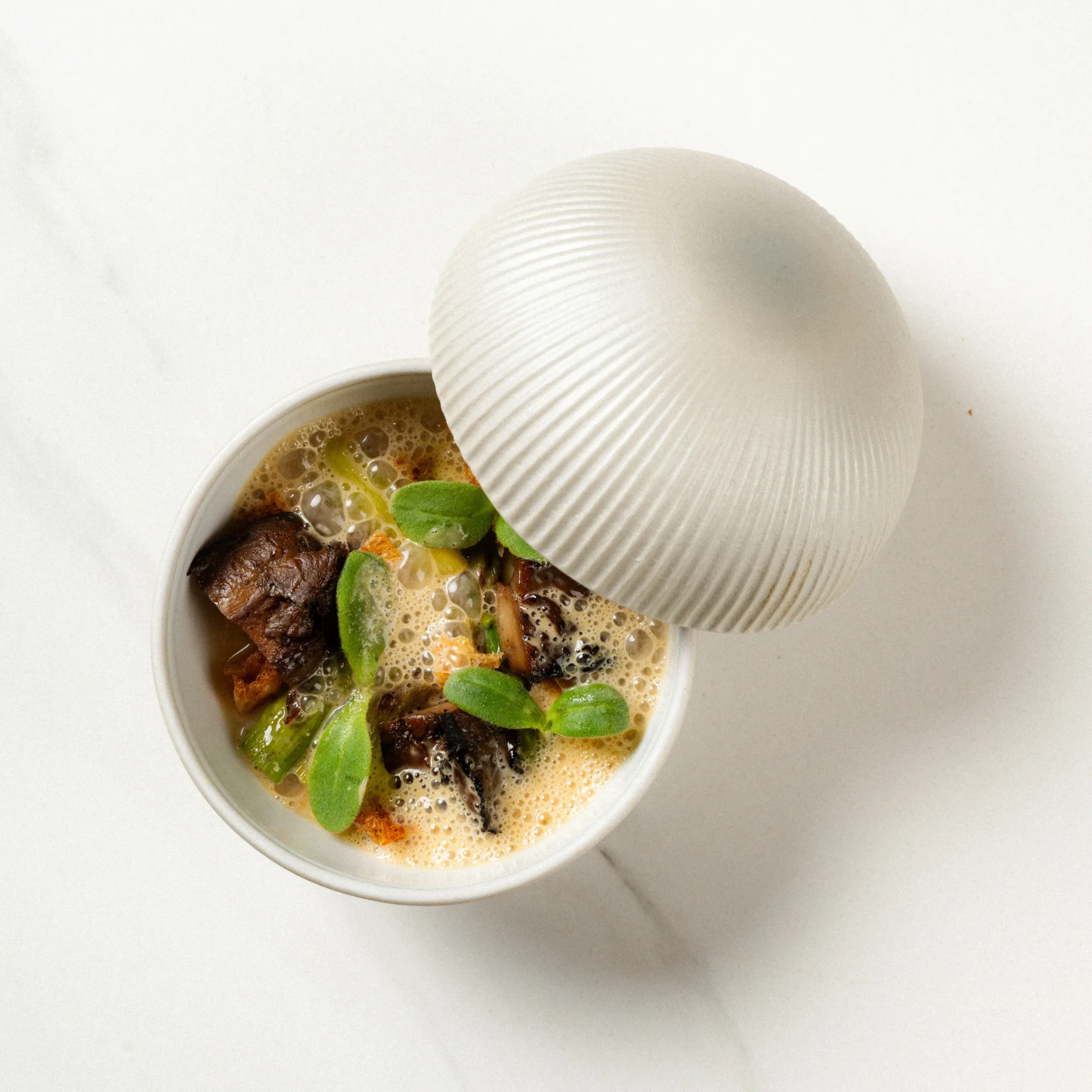 Top-down view of a white bowl with foamy broth, chunks of meat, and green herbs, partially covered by a white lid.