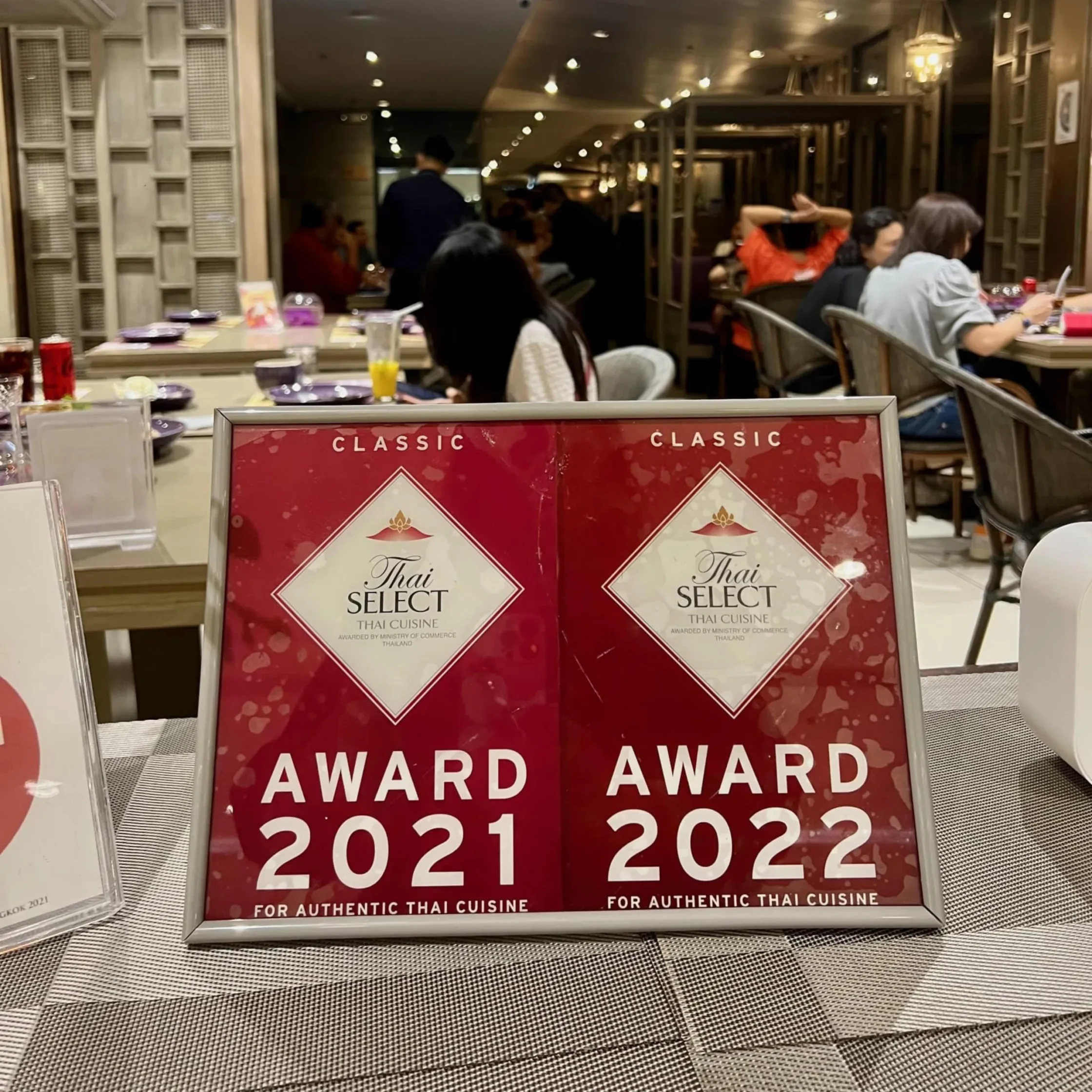 Red signage reading 'Thai Select' awards in a restaurant setting, with 'Award 2021 2022' visible on the card in the foreground.