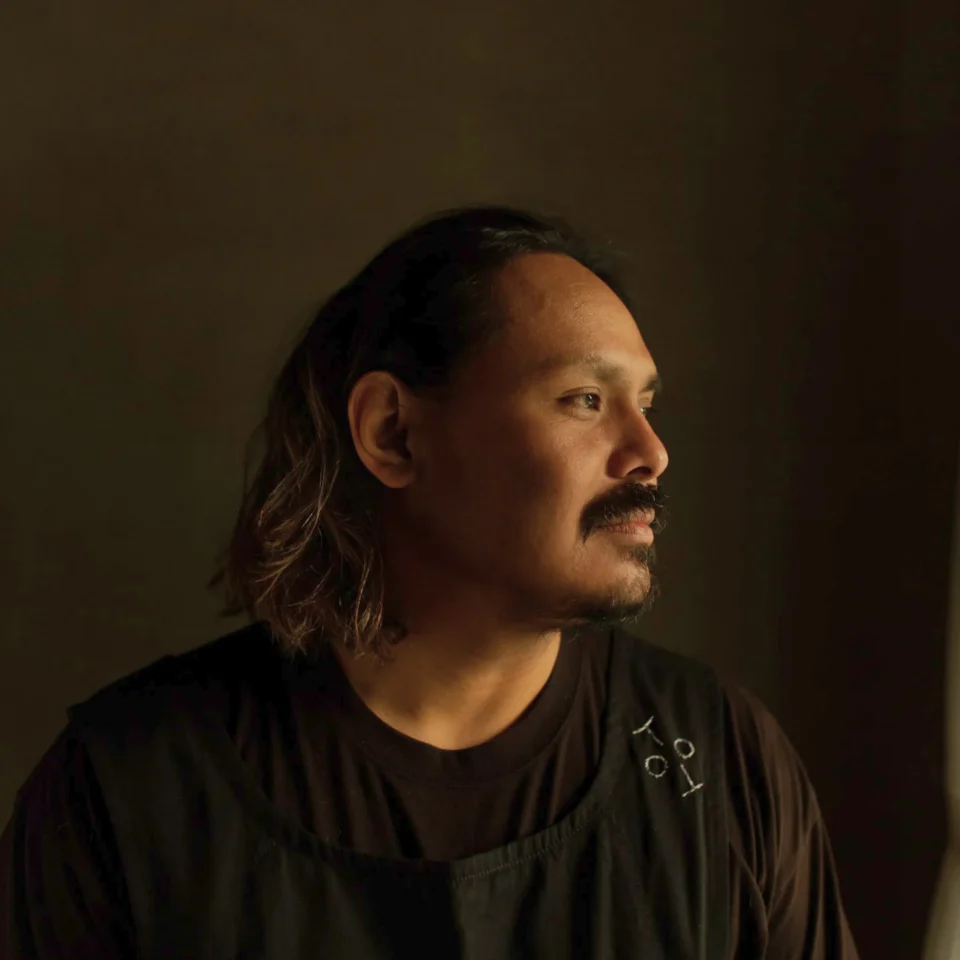 Side profile of a man with long hair and a mustache wearing a dark T-shirt against a dark background, dim lighting.