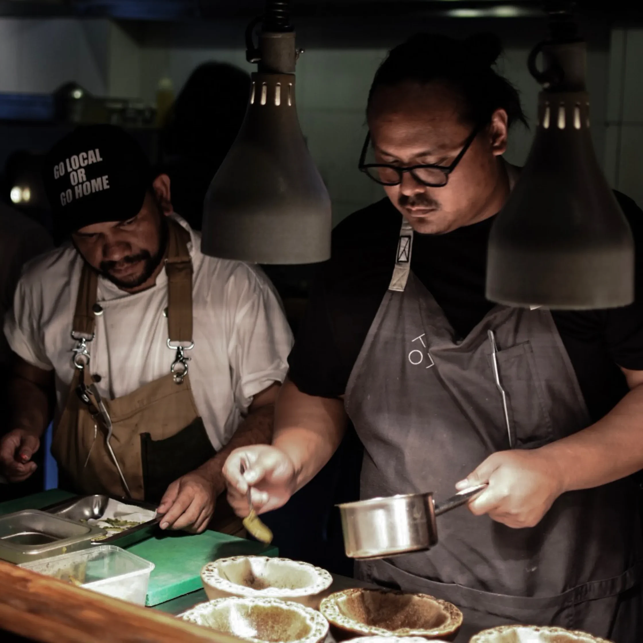 Two cooks in an dimly lit kitchen shape and warm tortillas on a griddle, assembling taco shells with toppings nearby.