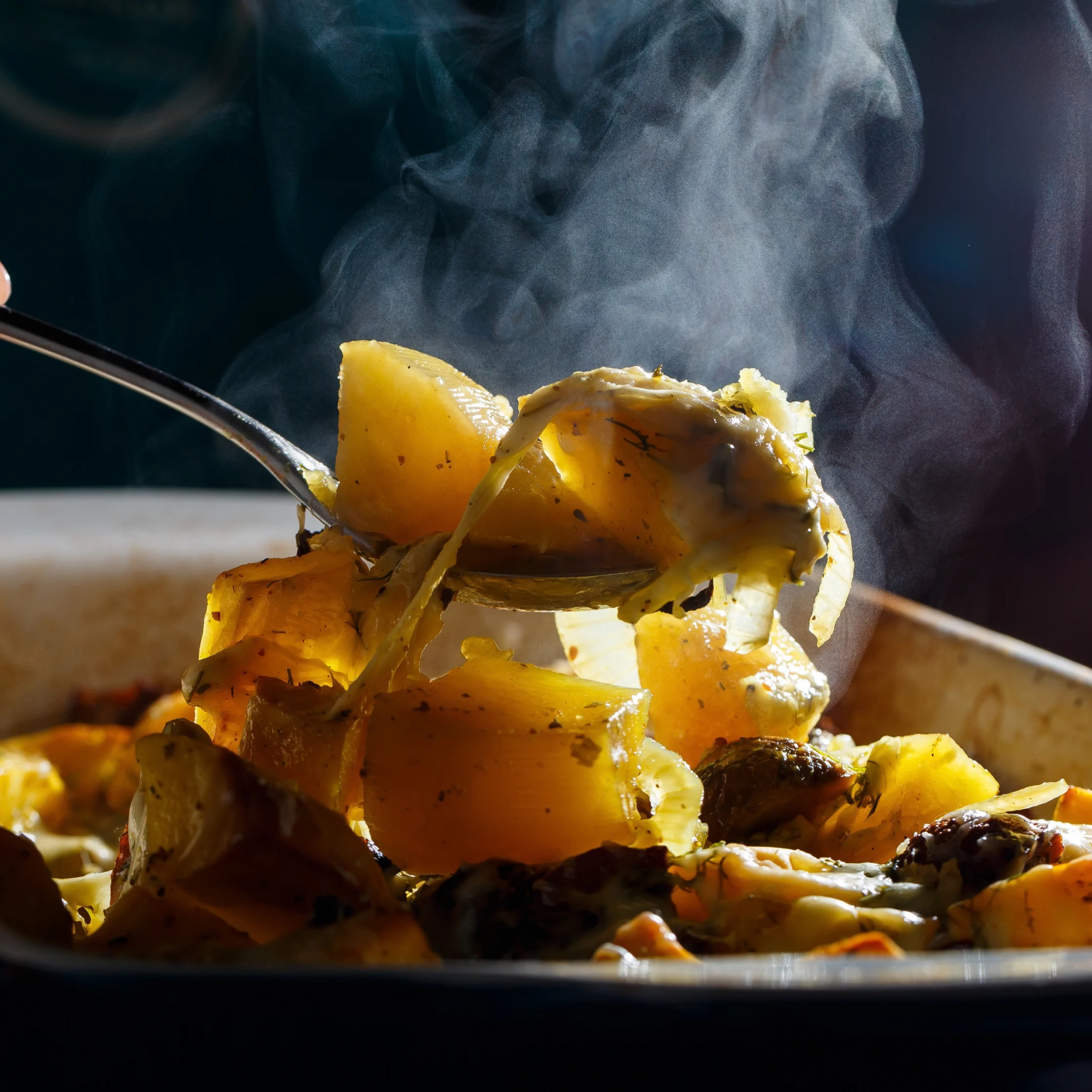 A spoon lifts tender golden potato chunks and vegetables from a pan, with steam curling upward; warm light evokes a cozy, freshly cooked meal.