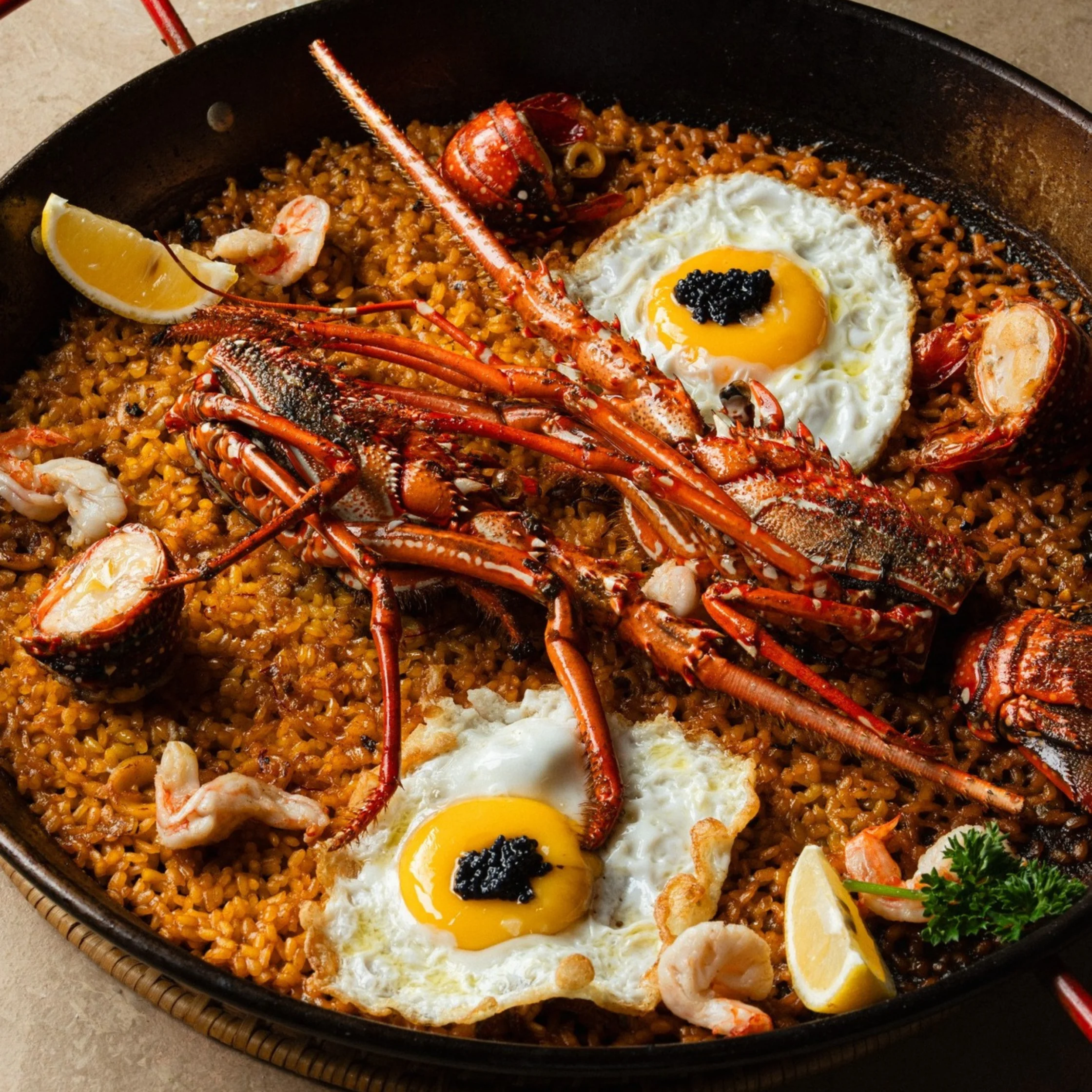 Rustic seafood paella in a large pan, topped with lobster, shrimp, squid, lemon wedges, parsley, and two sunny-side-up eggs for a festive meal.
