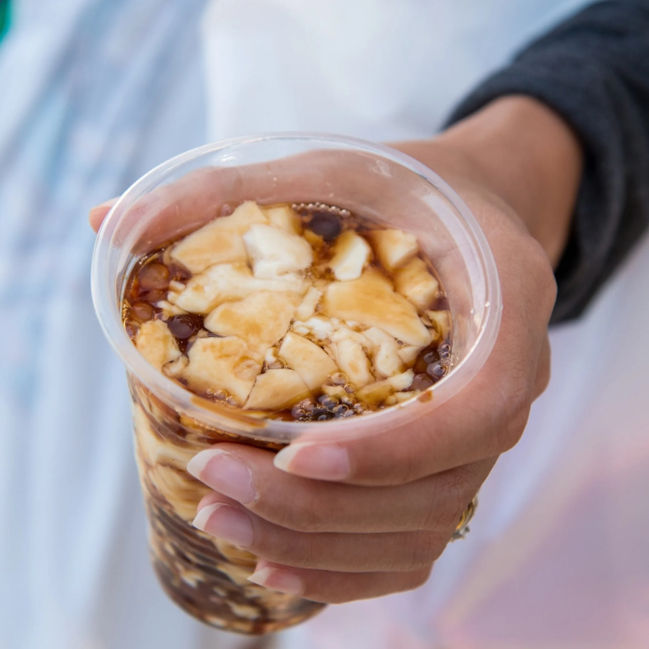 A hand presents a clear cup of iced coffee topped with diced banana and cream, with syrup and ice visible below; the mood is casual and inviting.