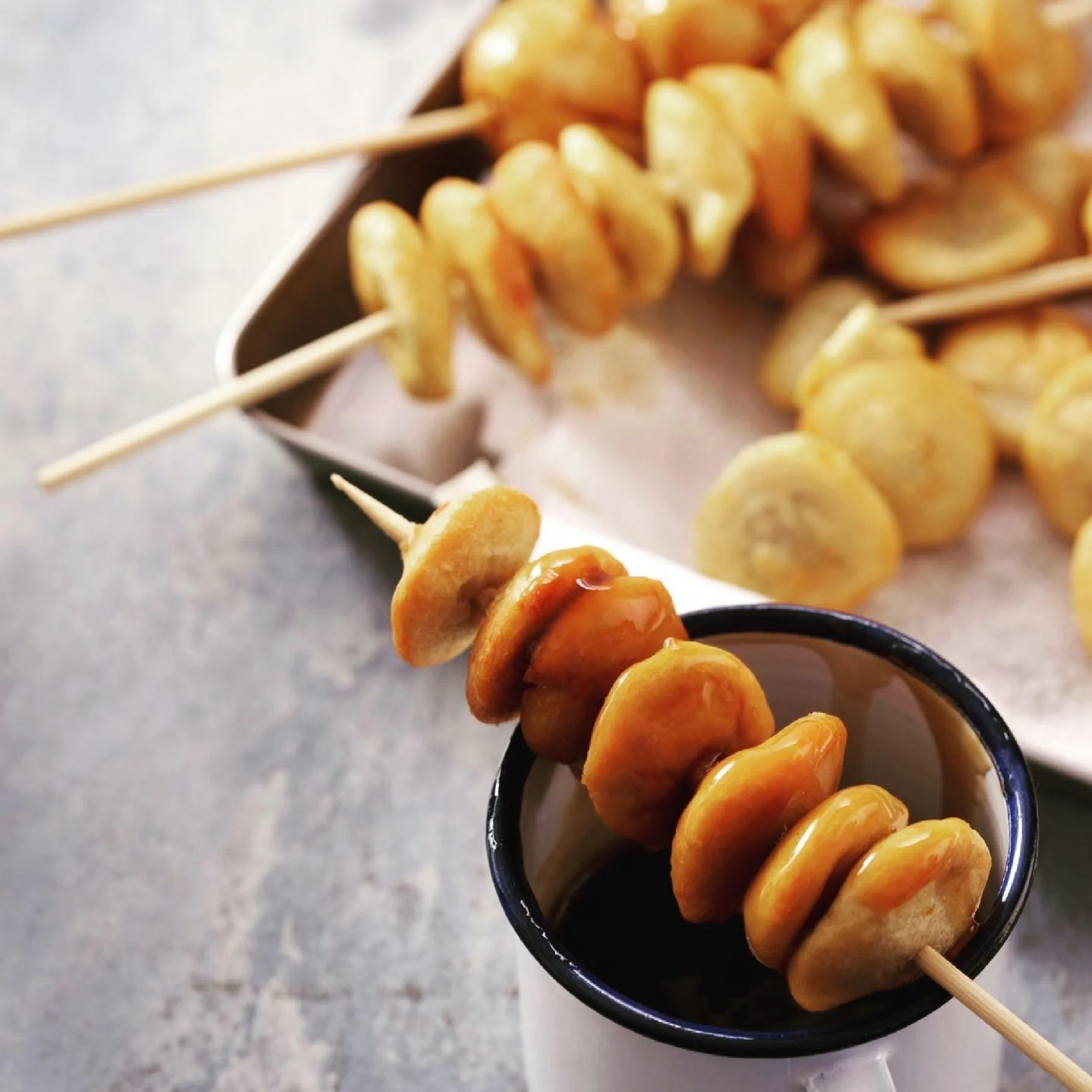 Golden spiral-cut potato skewers and round fried potato pieces are dipped in dark sauce in a black cup, with more skewers blurred behind; warm, appetizing street-food scene.