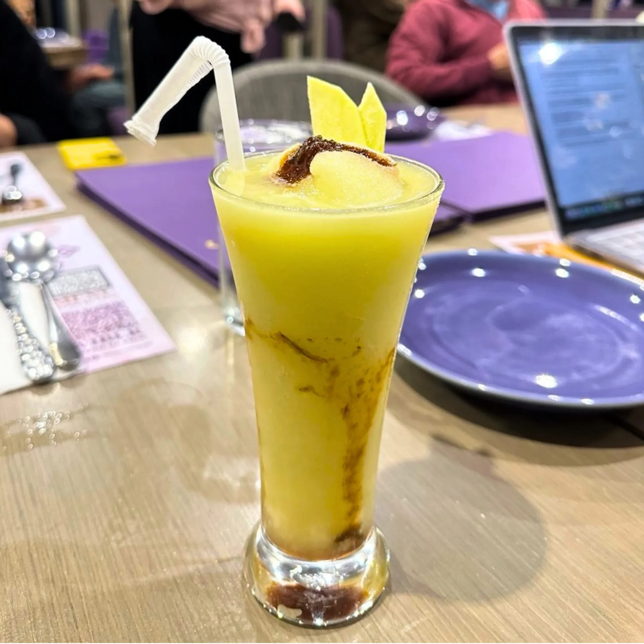 Tall glass of yellow tropical drink with pineapple wedges garnish and a bendy white straw on a cafe table, laptop blurred in background