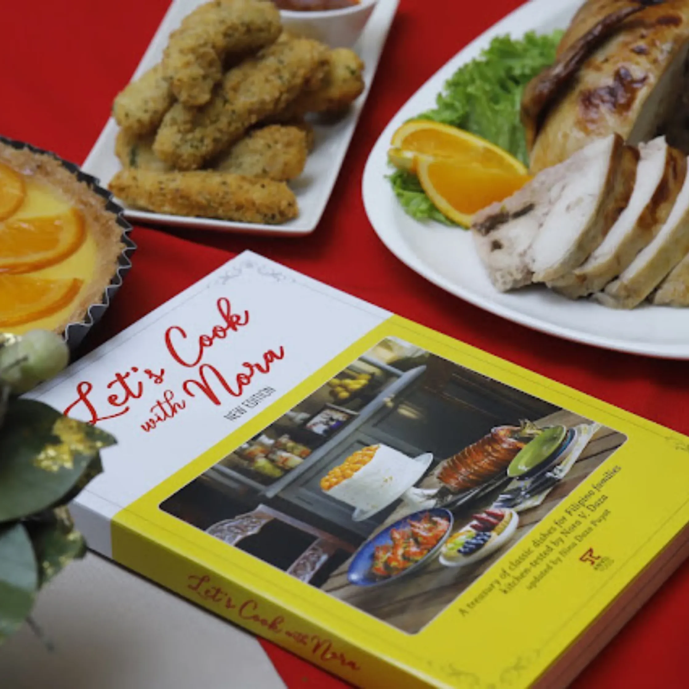Assorted fried appetizers on a white plate with a dipping cup, orange slice garnish, and a loaf on a red table.
