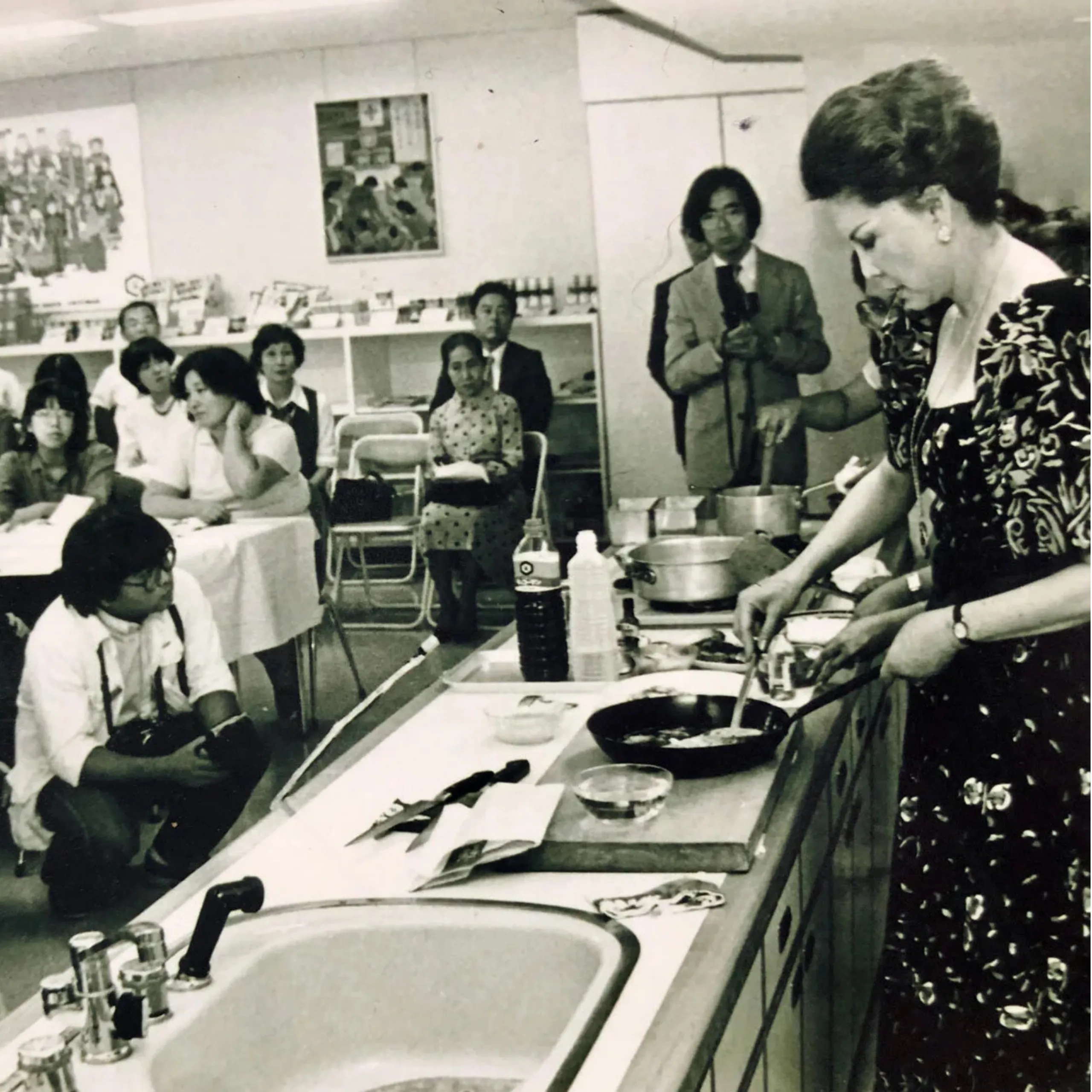 A woman in a patterned dress demonstrates cooking at a counter while an audience watches in a classroom or school cafeteria.