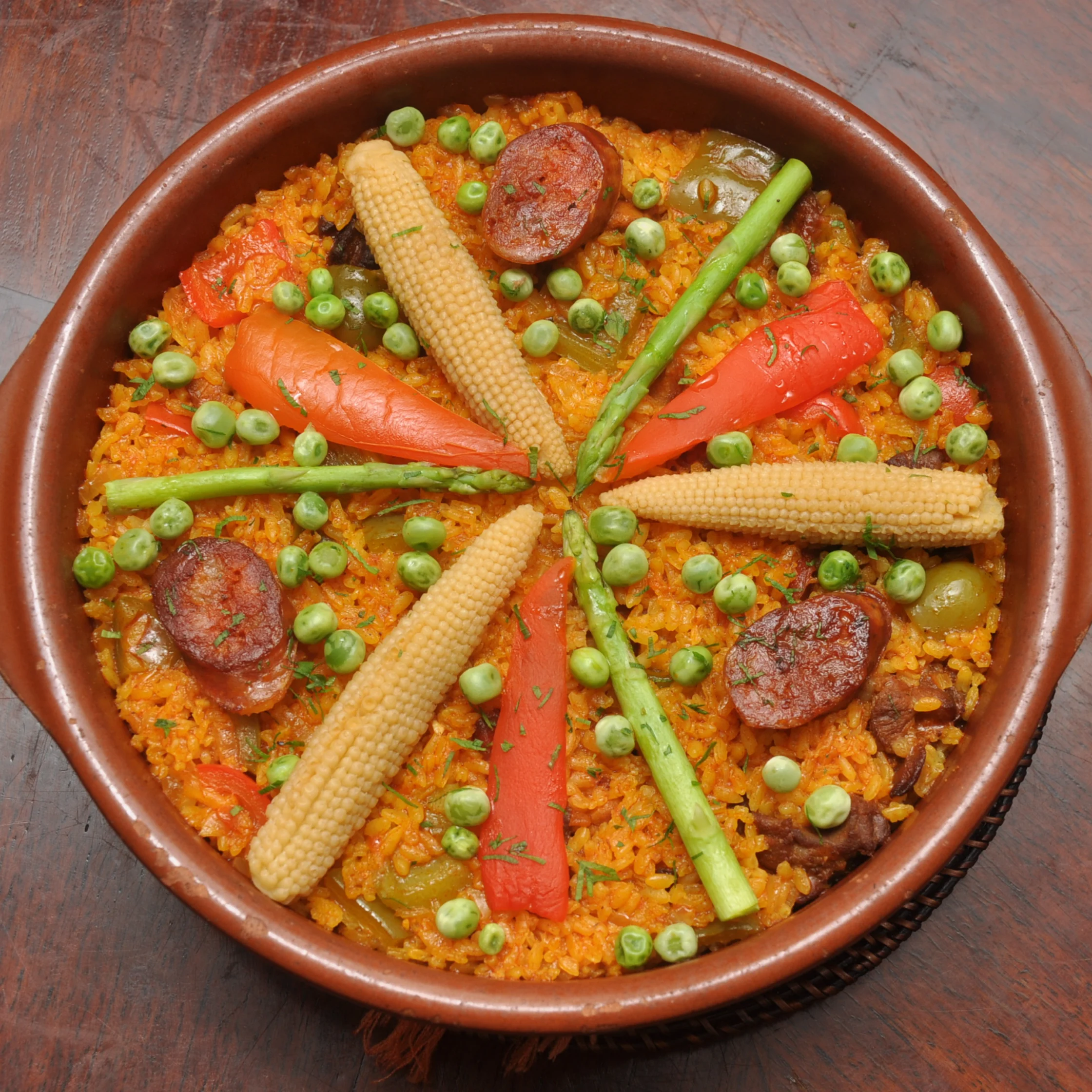 Overhead view of colorful vegetable paella in a rustic brown dish, topped with peas, baby corn, red peppers, asparagus, and chorizo.