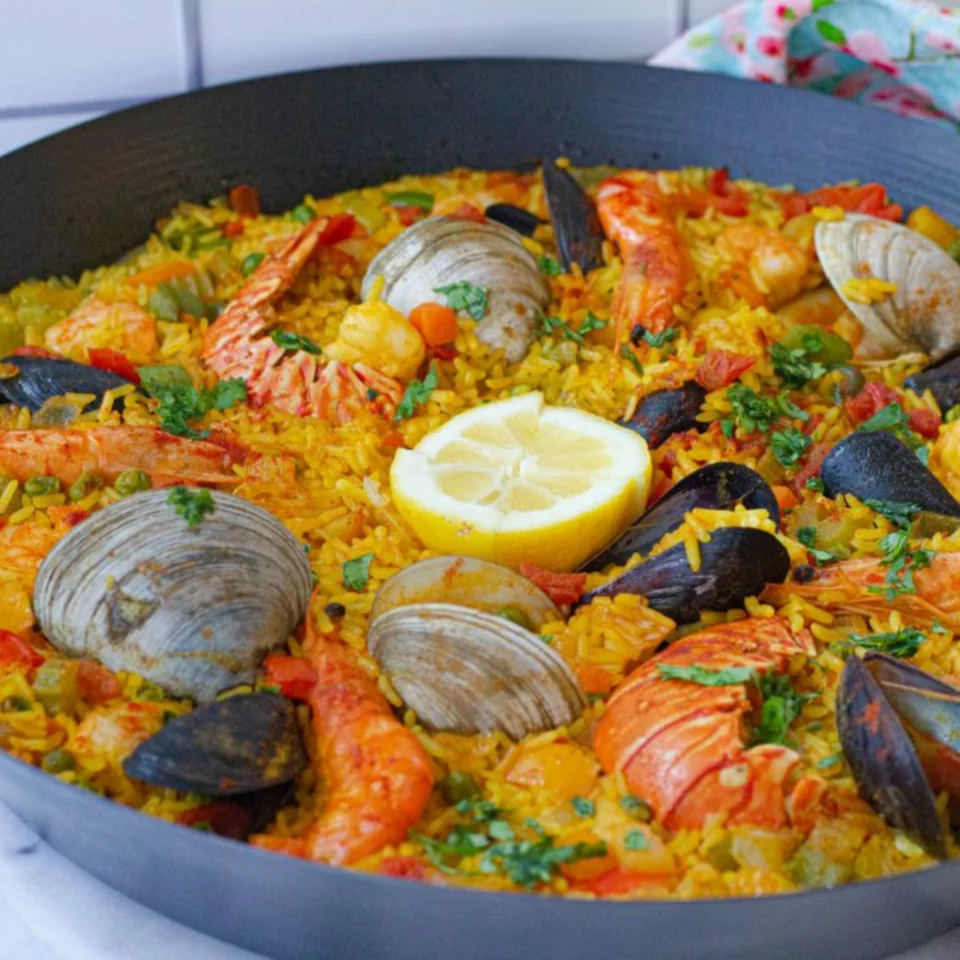 Colorful seafood paella in a black pan, topped with shrimp, clams, mussels, lobster, herbs, and a lemon half; warm, inviting dish.