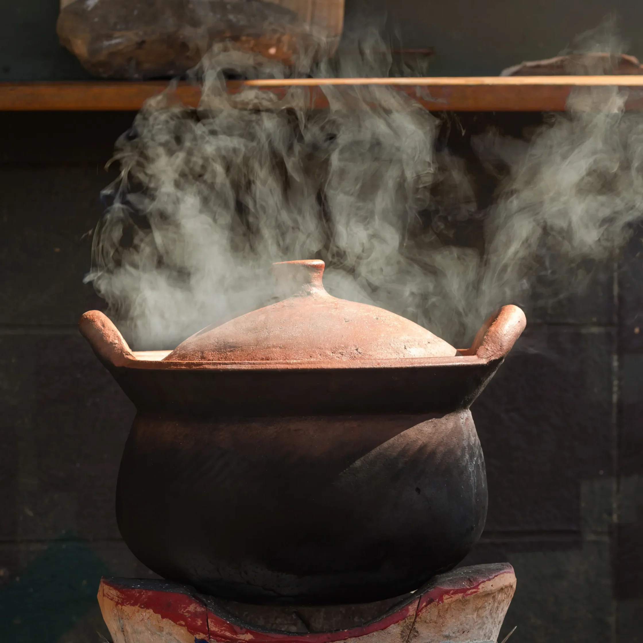 Large rustic clay pot with a red rim, releasing steam in a dim kitchen setting.