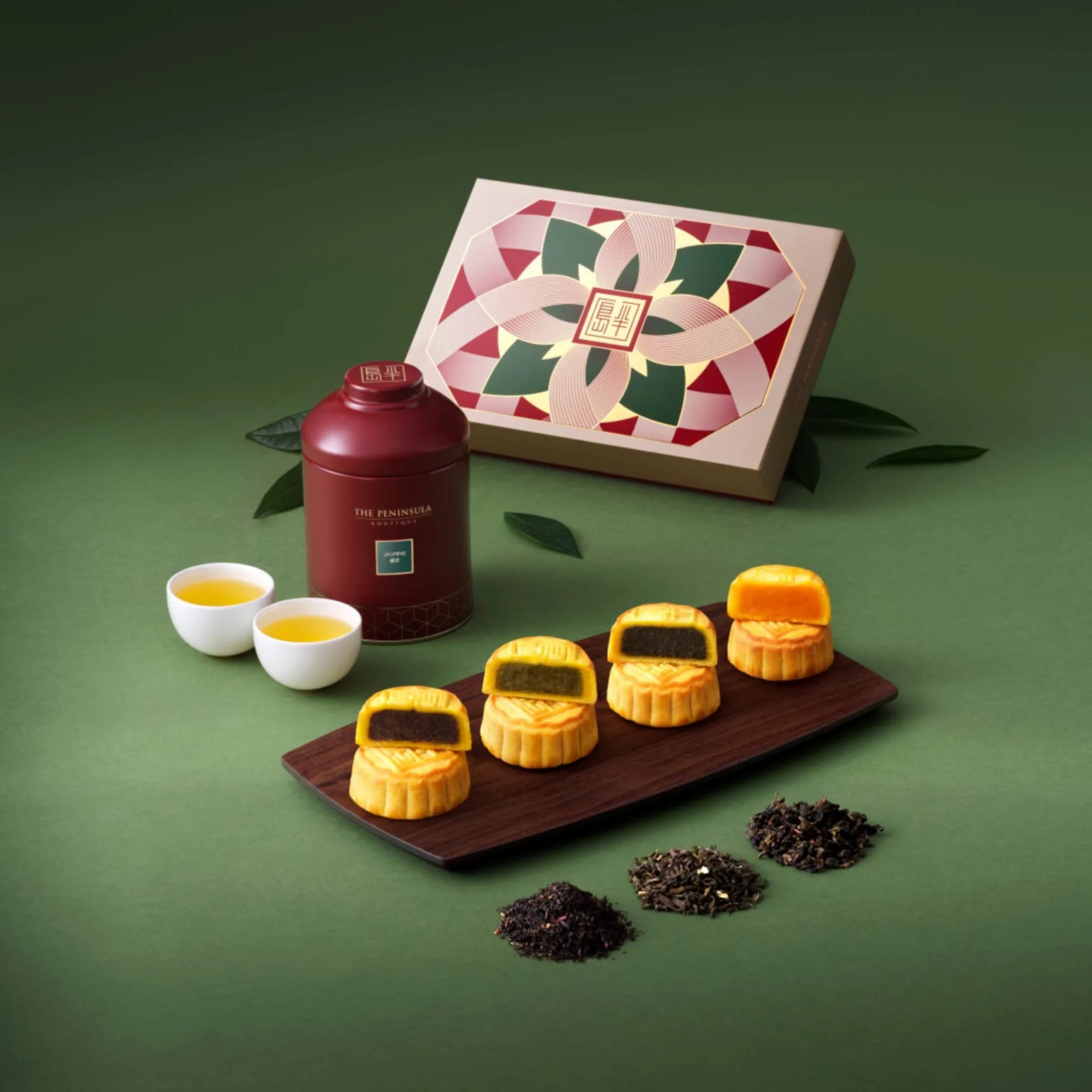 Elegant tea gift set on a green backdrop: four golden mooncakes, a red tea tin, teacups, loose tea leaves, and a patterned box convey warmth.