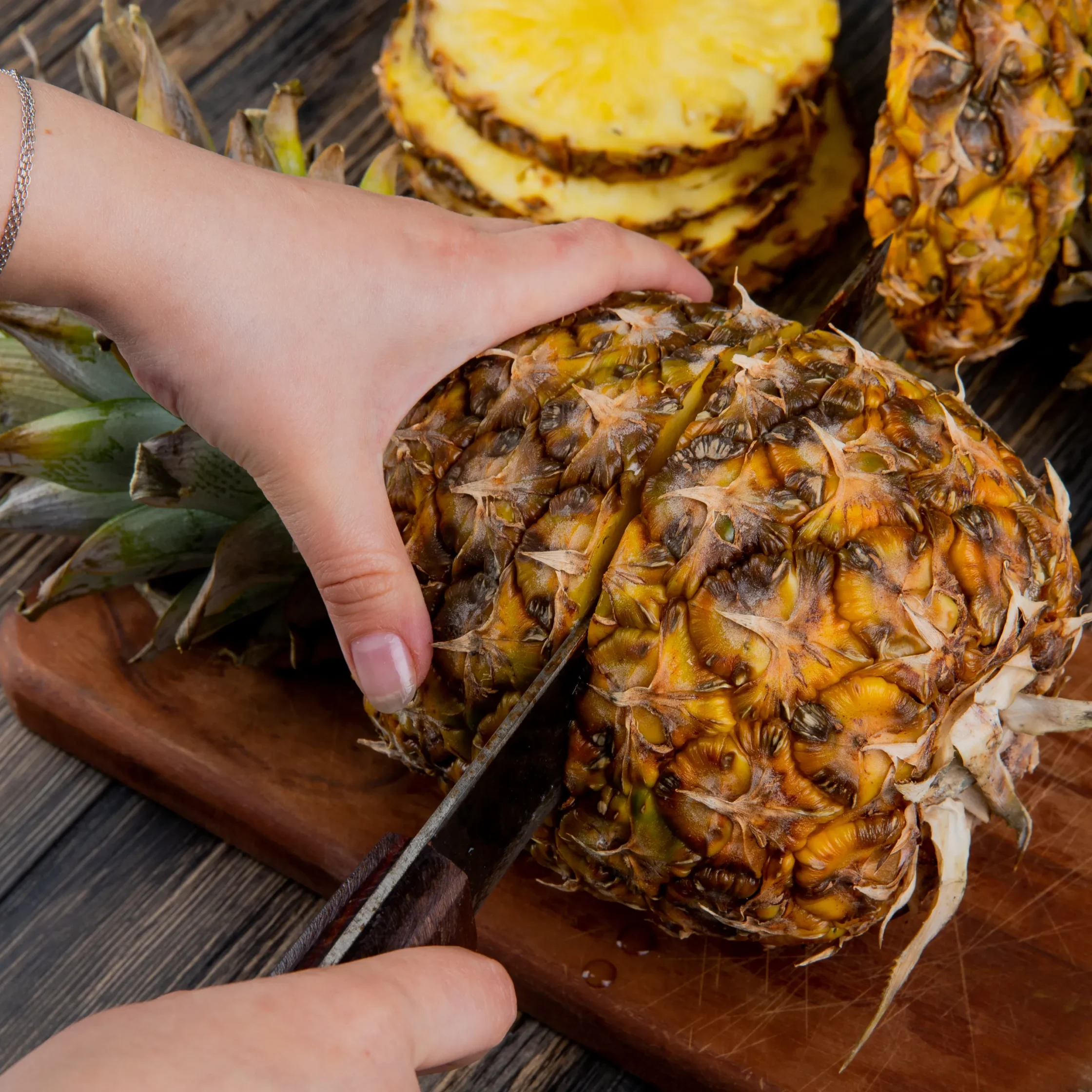 Close-up of a hand slicing a pineapple on a wooden cutting board.