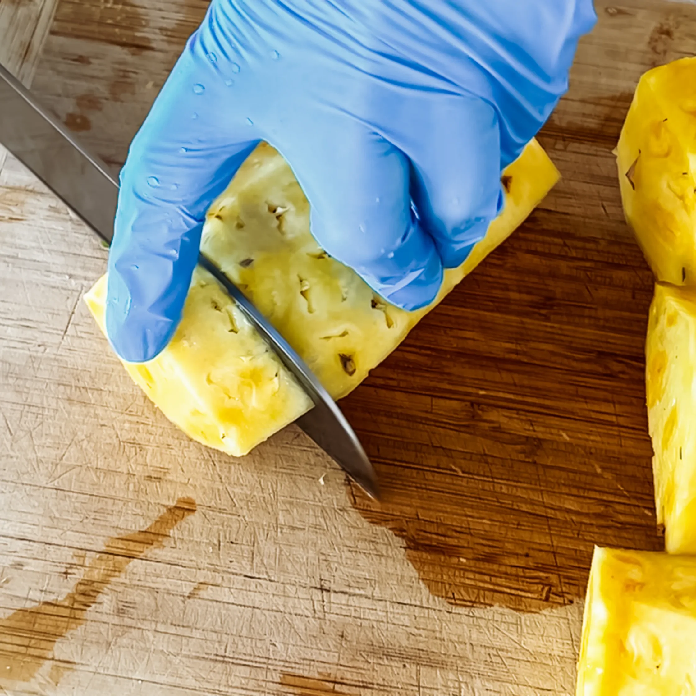 Close-up of a blue disposable glove on a hand over a yellow vegetable on a wooden cutting board with a knife nearby.