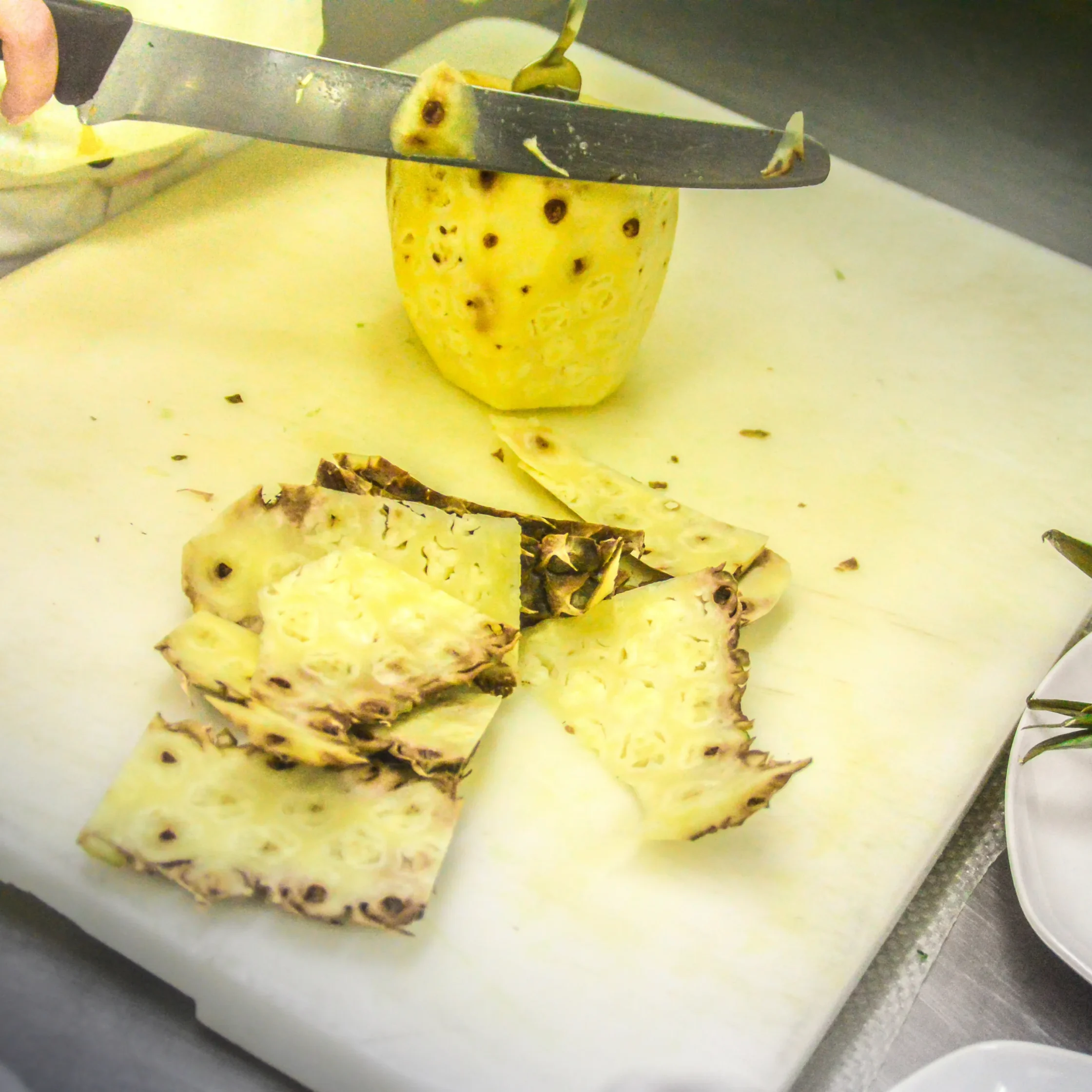 Close-up of a chef's knife slicing a pineapple on a cutting board, with pineapple pieces and scraps nearby.