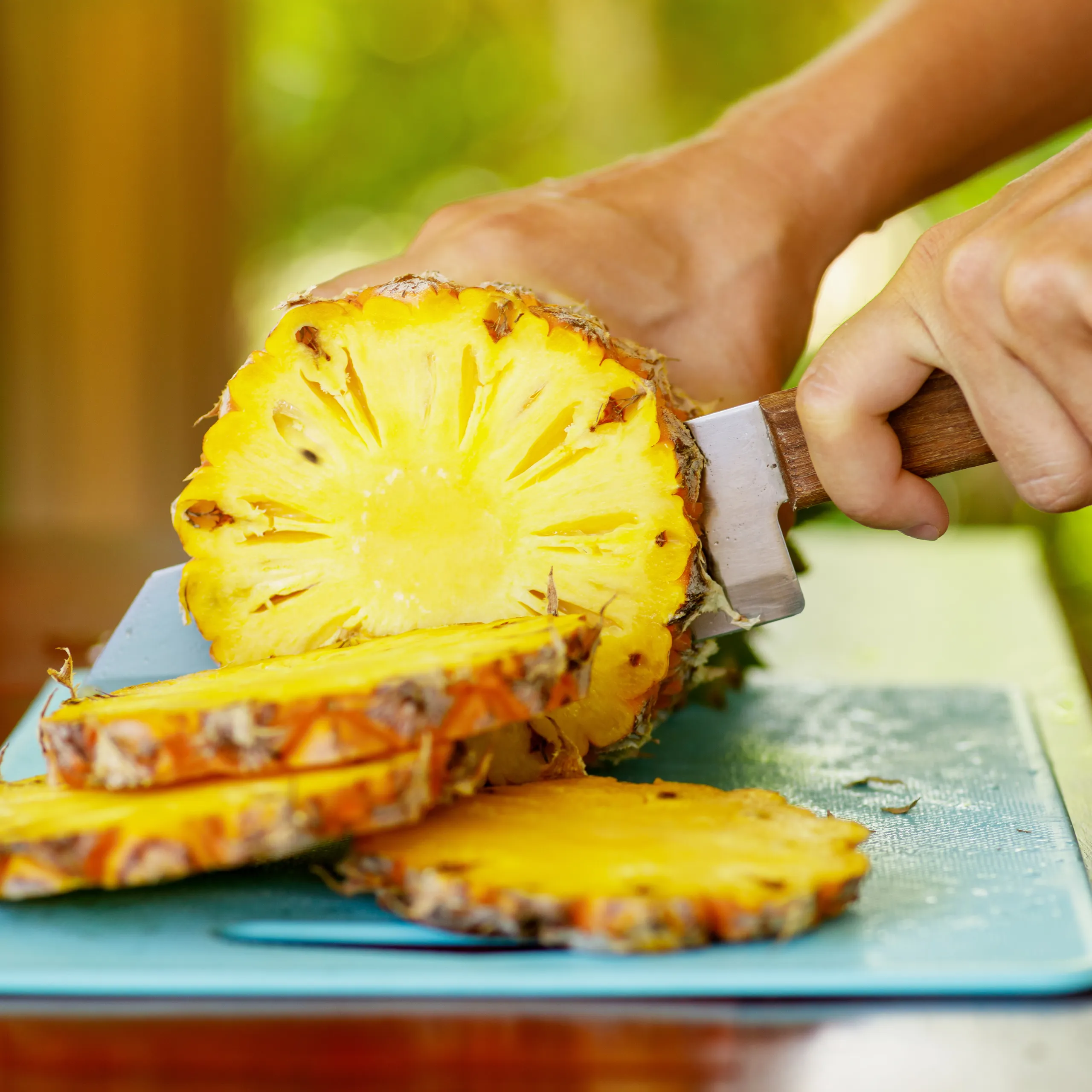 Close-up of a hand slicing a pineapple with a knife on a turquoise plate outdoors, pineapple rings piling up as they are cut.