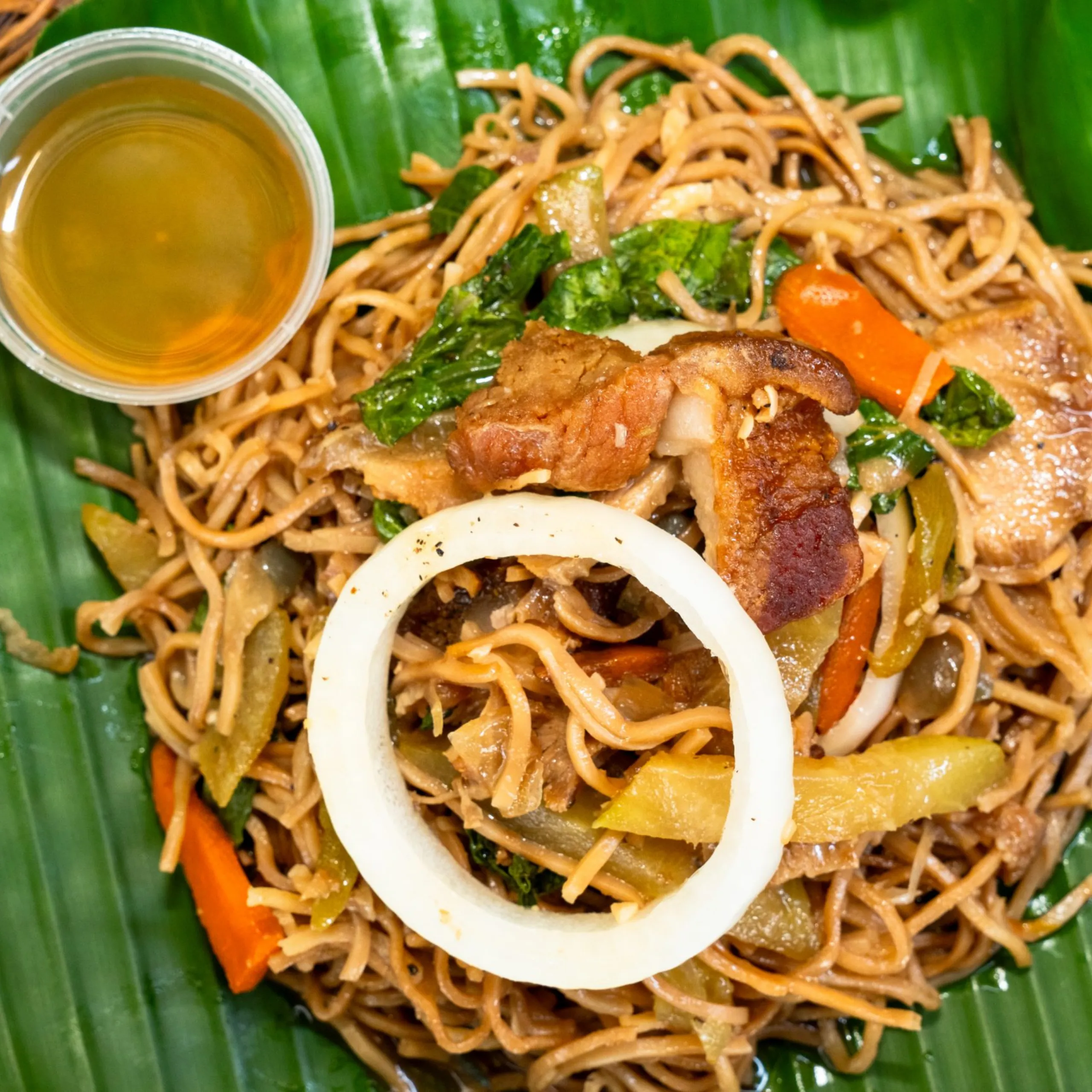 Pad Thai–style noodles with vegetables on a banana leaf, served with a cup of sauce.