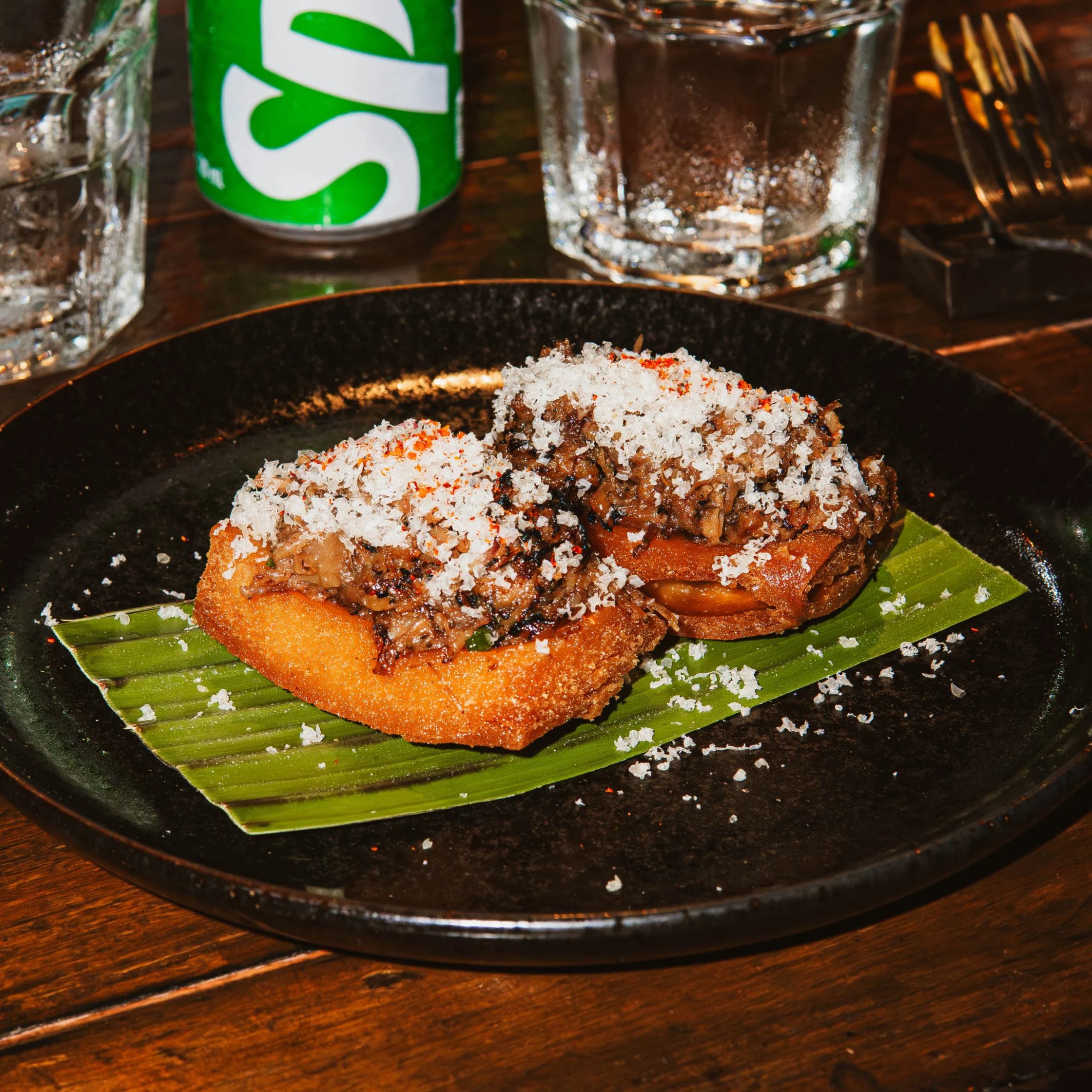 Close-up of toasted bread topped with shredded meat and large flaky salt on a banana leaf, with a glass and green can in the background.