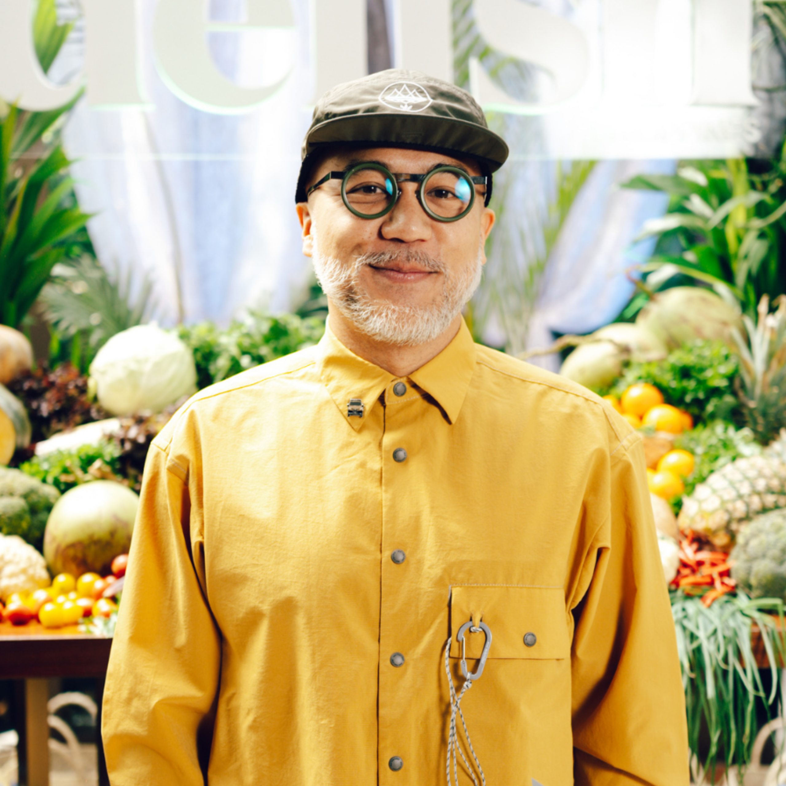 Man wearing a mustard-yellow shirt and round green glasses, standing at a bright produce market with fruits in the background.