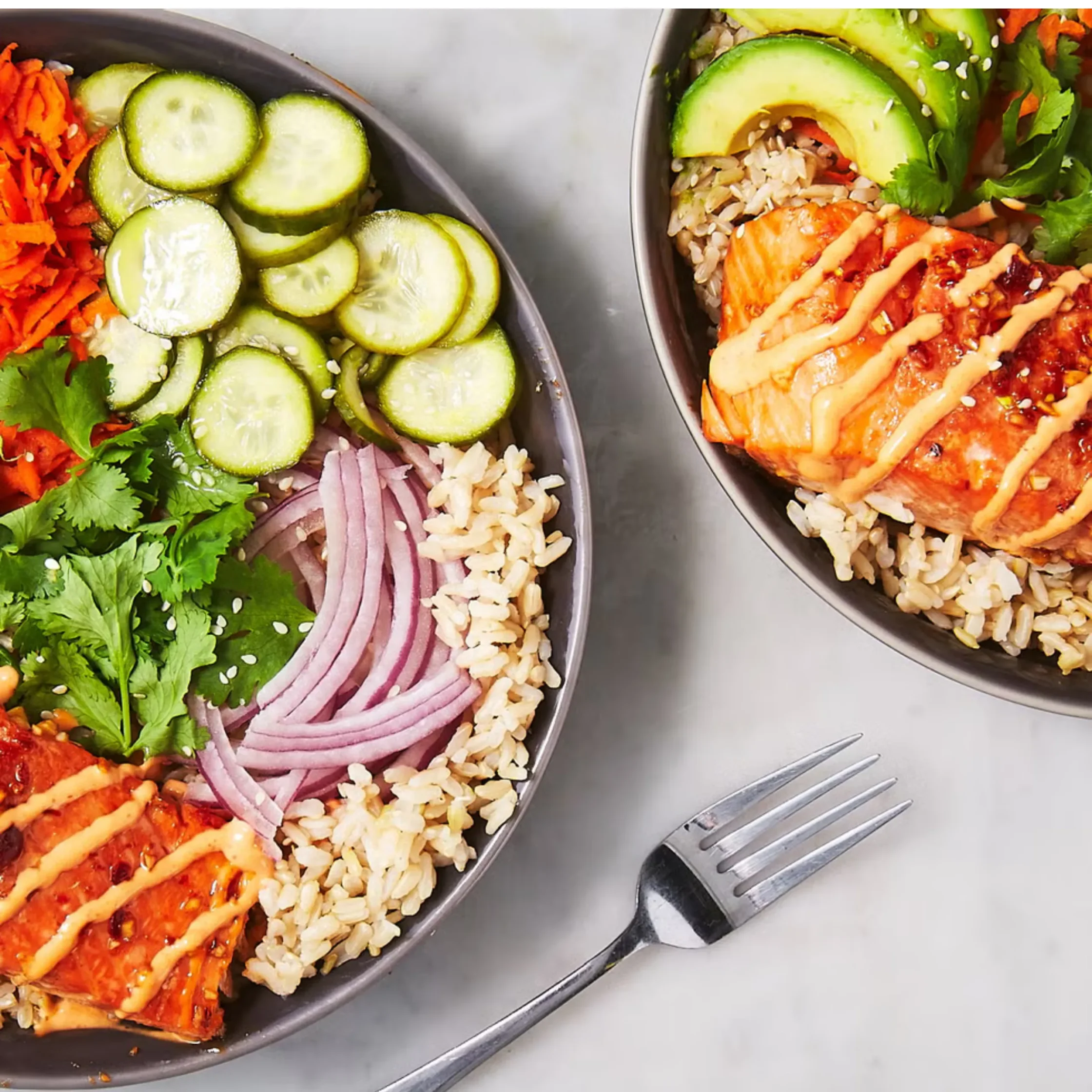 Top-down view of two colorful rice bowls: cucumber slices, shredded carrot, cilantro, ham, chicken with sauce, rice, and avocado garnishes with sesame seeds.